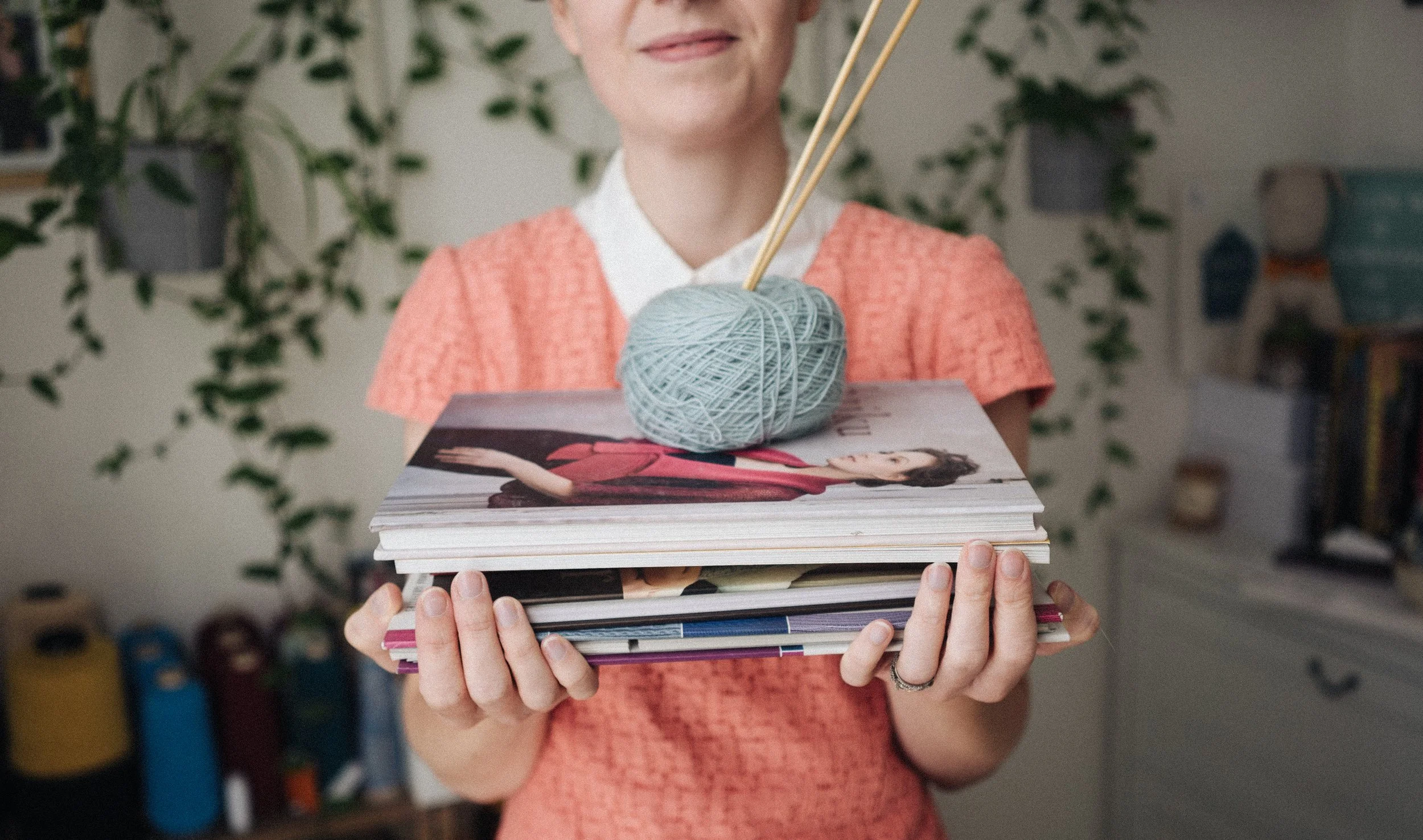 Knitting Teacher Alice holds a stack of knitting pattern books with a ball of blue yarn and wooden needles sitting on the top