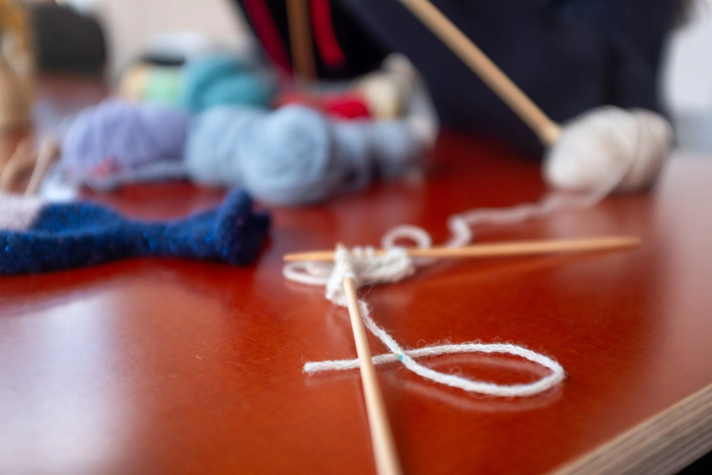 Close-up of a brown table with various balls of yarn, knitting needles, and white thread scattered across the surface.