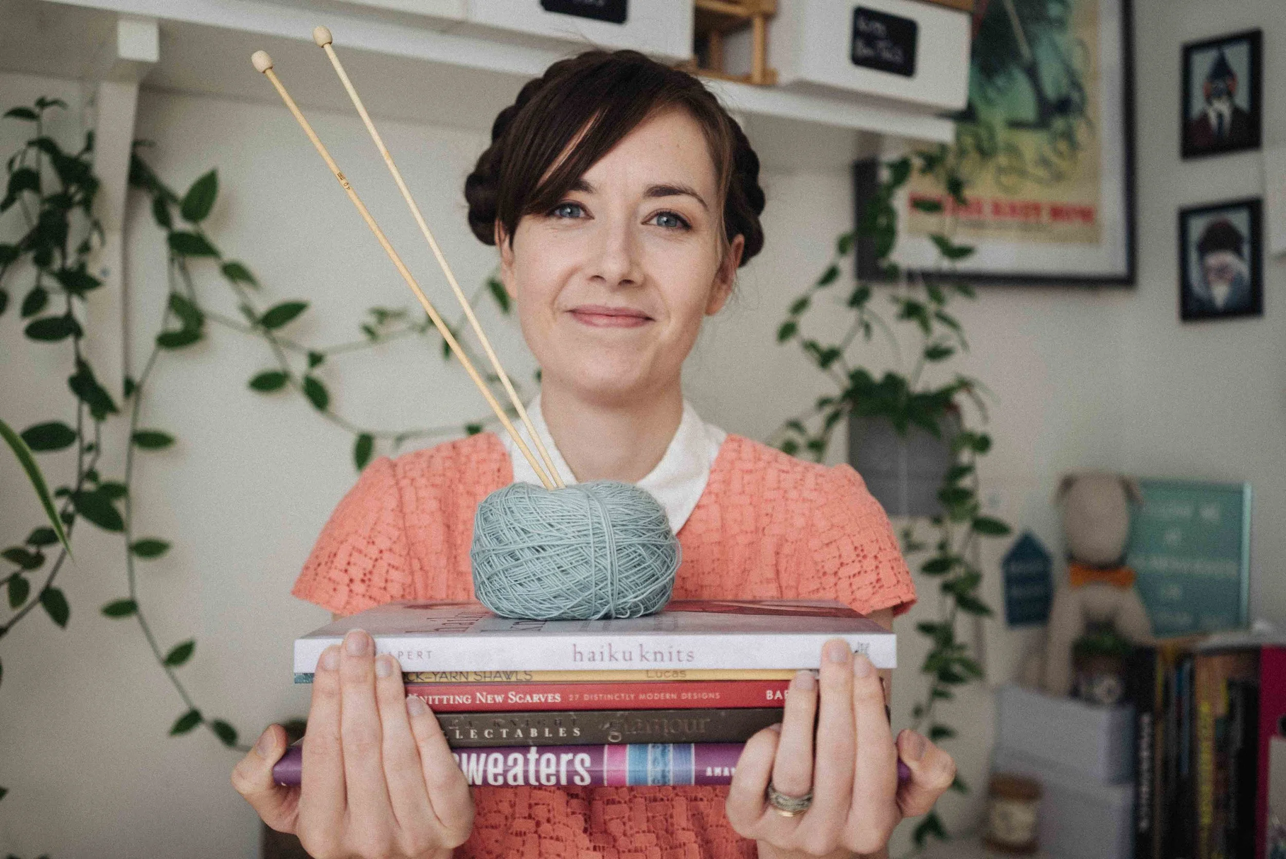 A woman with short dark hair and a friendly smile holds a stack of books, with a ball of gray yarn and knitting needles on top, in a cozy room decorated with plants and framed pictures.