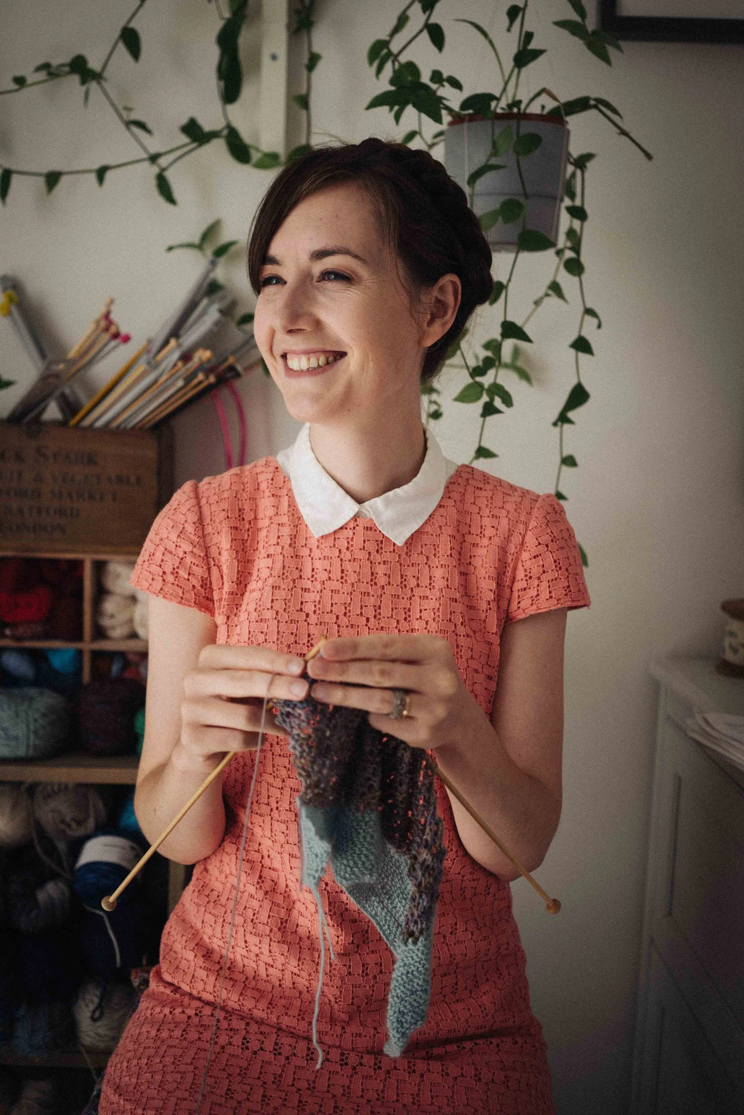 Woman smiling while knitting with yarn and knitting needles, wearing a coral lace dress with a white collar, in a room with shelves of yarn and yarn supplies in the background.