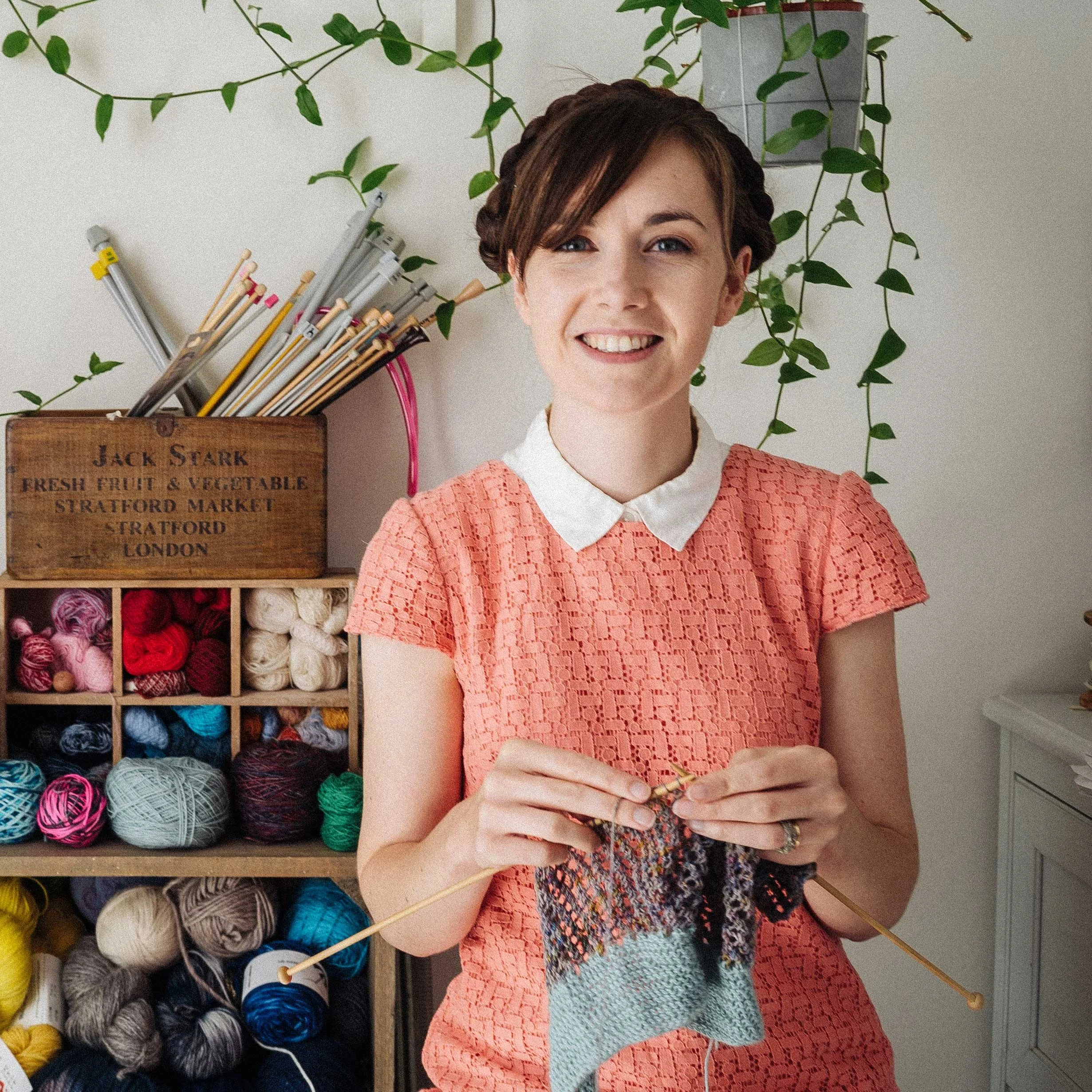 Alice a white female knitting teacher sits knitting and smiling at the camera in front of wooden boxes of yarn