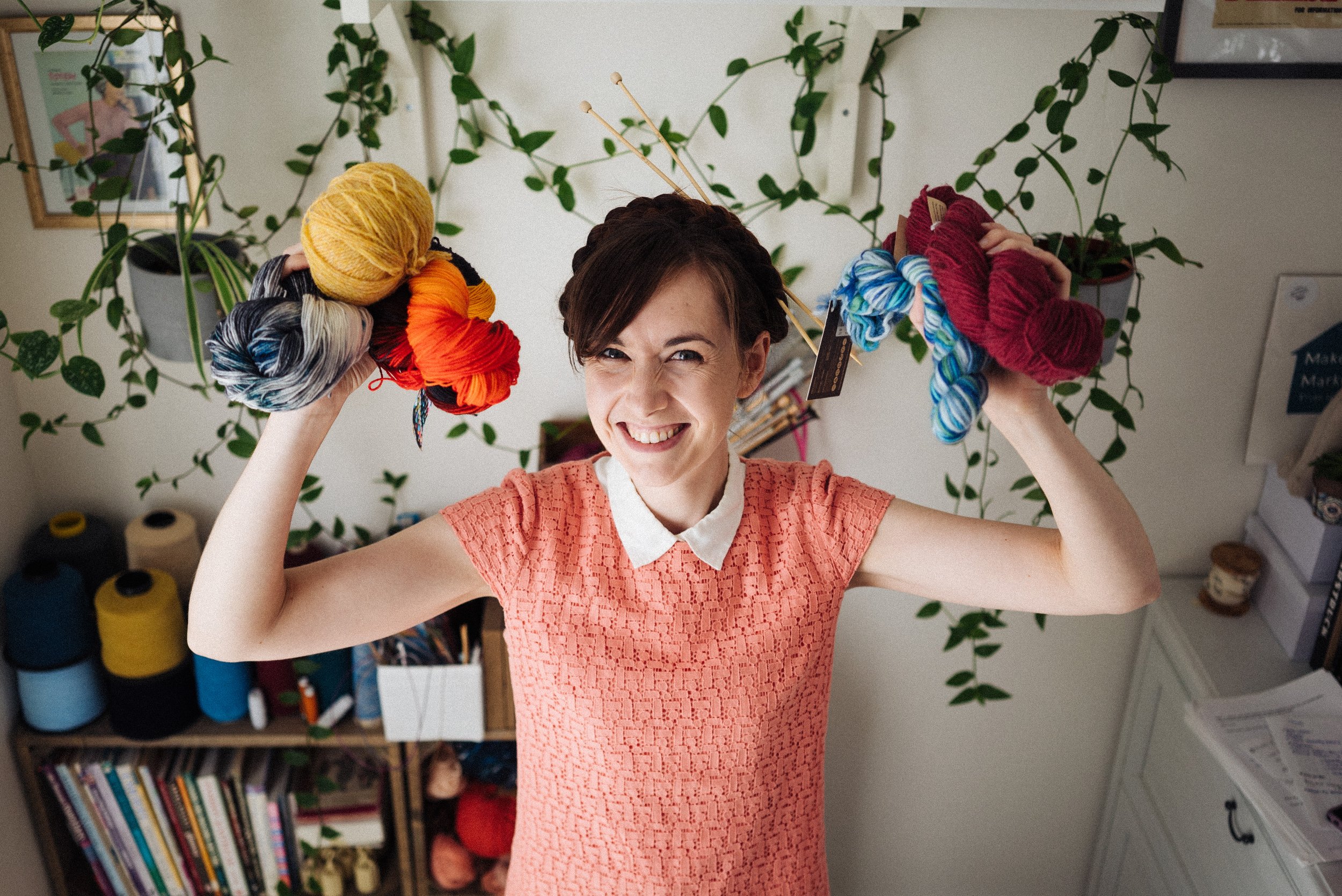 Alice a white female knitting teacher holds skeins of yarn either side of her head