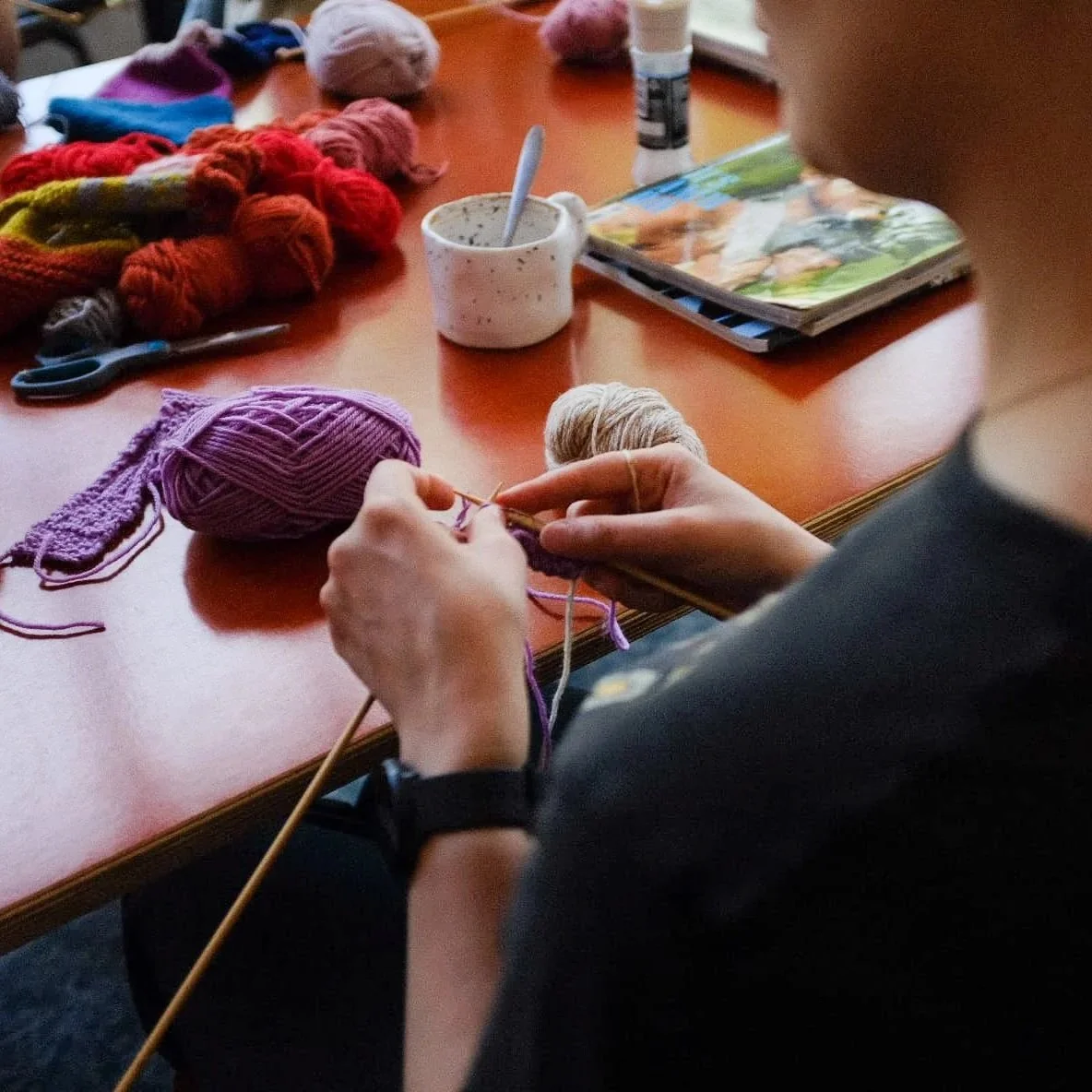 White, female hands knit with purple yarn and wooden needles at a cafe table