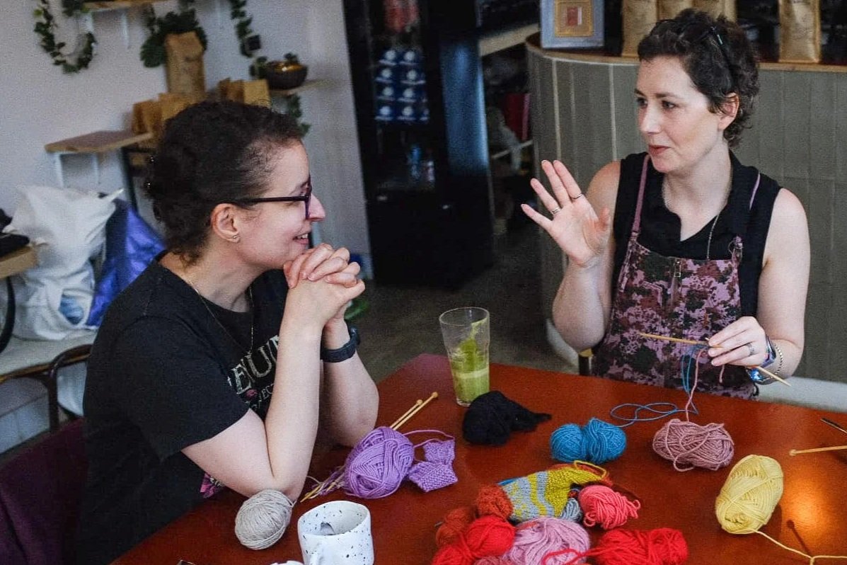 Two women are sitting at a table with yarn and knitting needles, engaging in conversation. One woman is smiling with her hands clasped, while the other is gesturing with her hand. The table has various balls of colored yarn, a cup, and knitting projects. The background includes shelves and decorative plants.