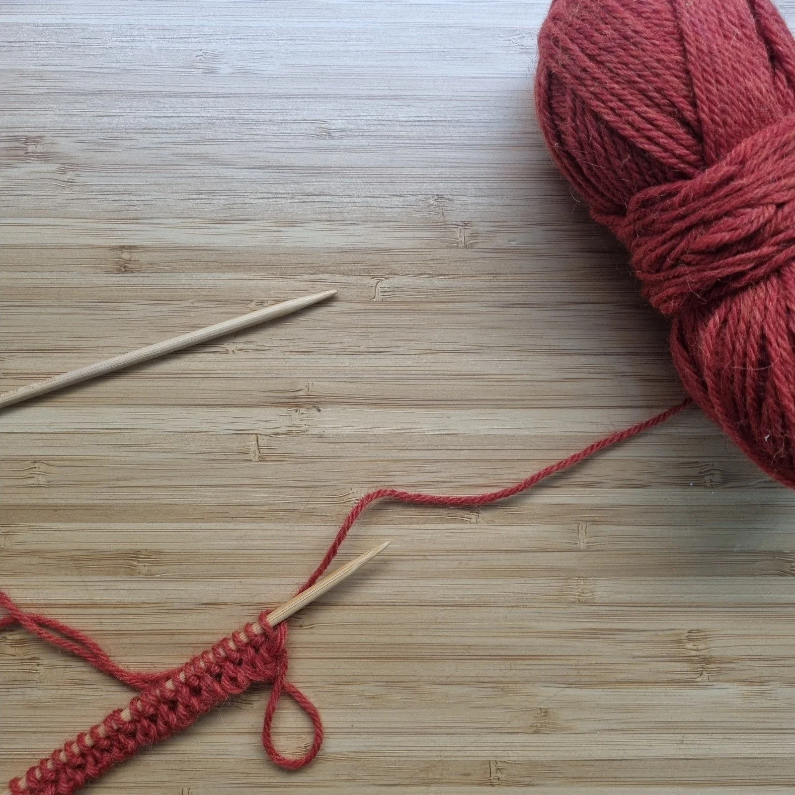 Two wooden knitting needles lay on a wooden surface with orange yarn and knitting on one needle