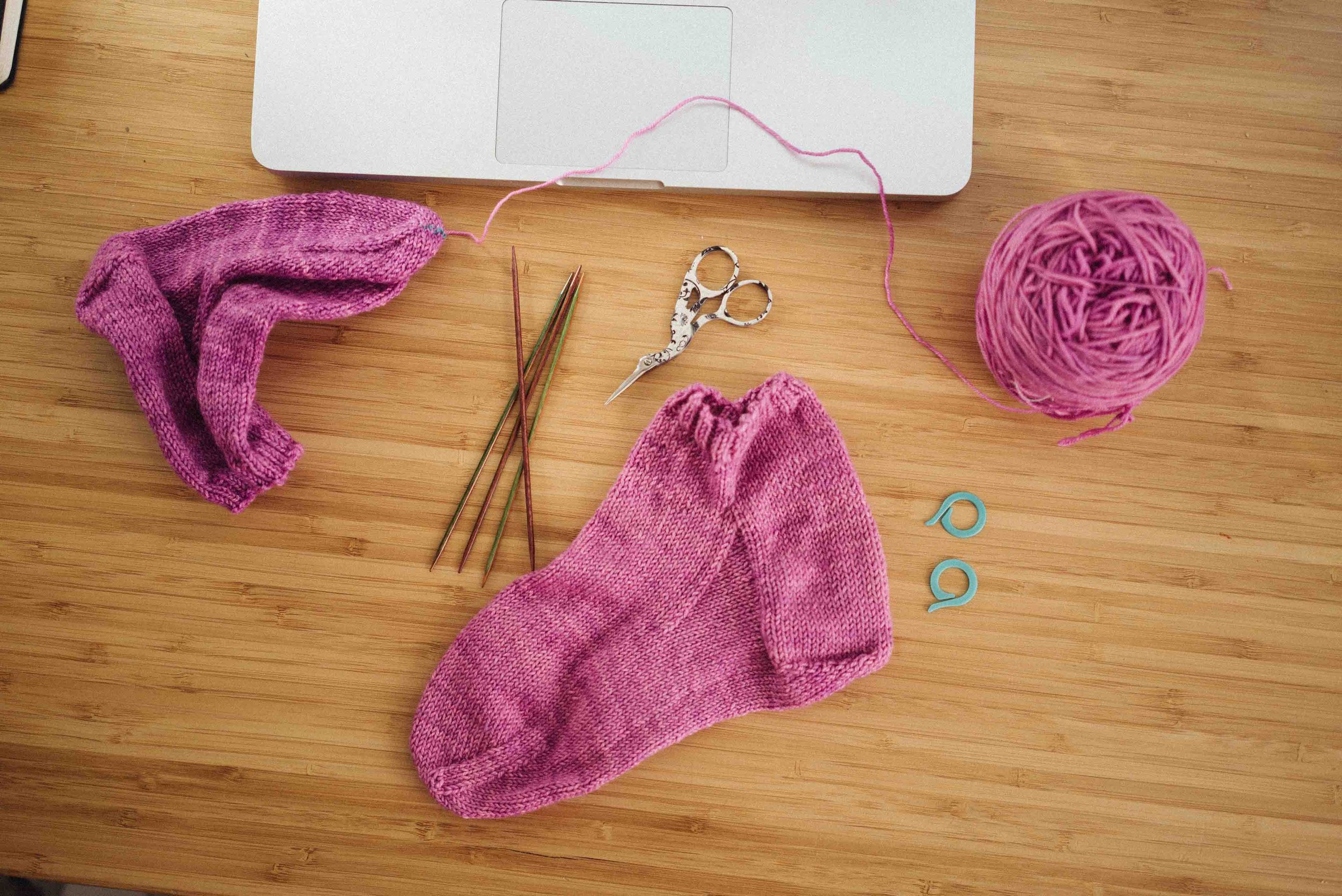 A knitting project in progress with pink yarn, partially knitted pink socks, knitting needles, scissors, and a ball of pink yarn on a wooden surface, with a silver laptop in the background.