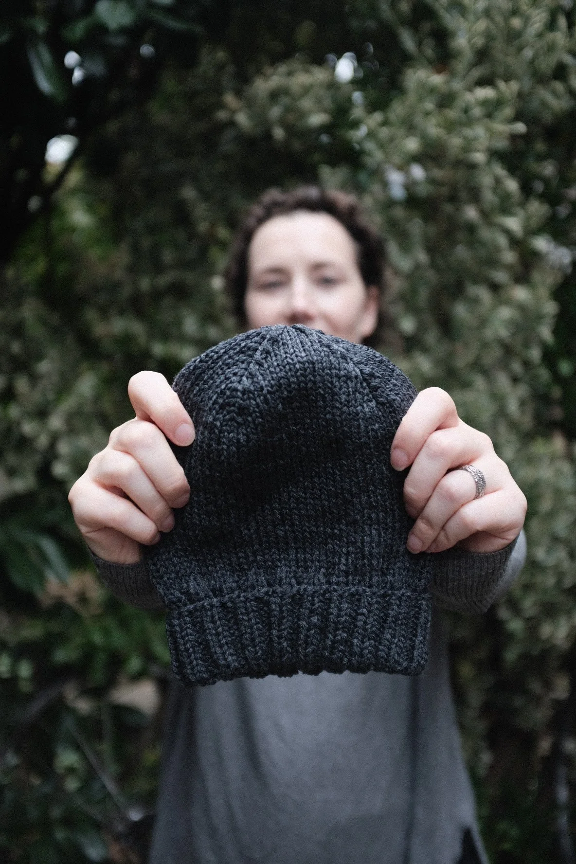 A person holding a dark gray knit beanie in front of their face outdoors, with greenery in the background.