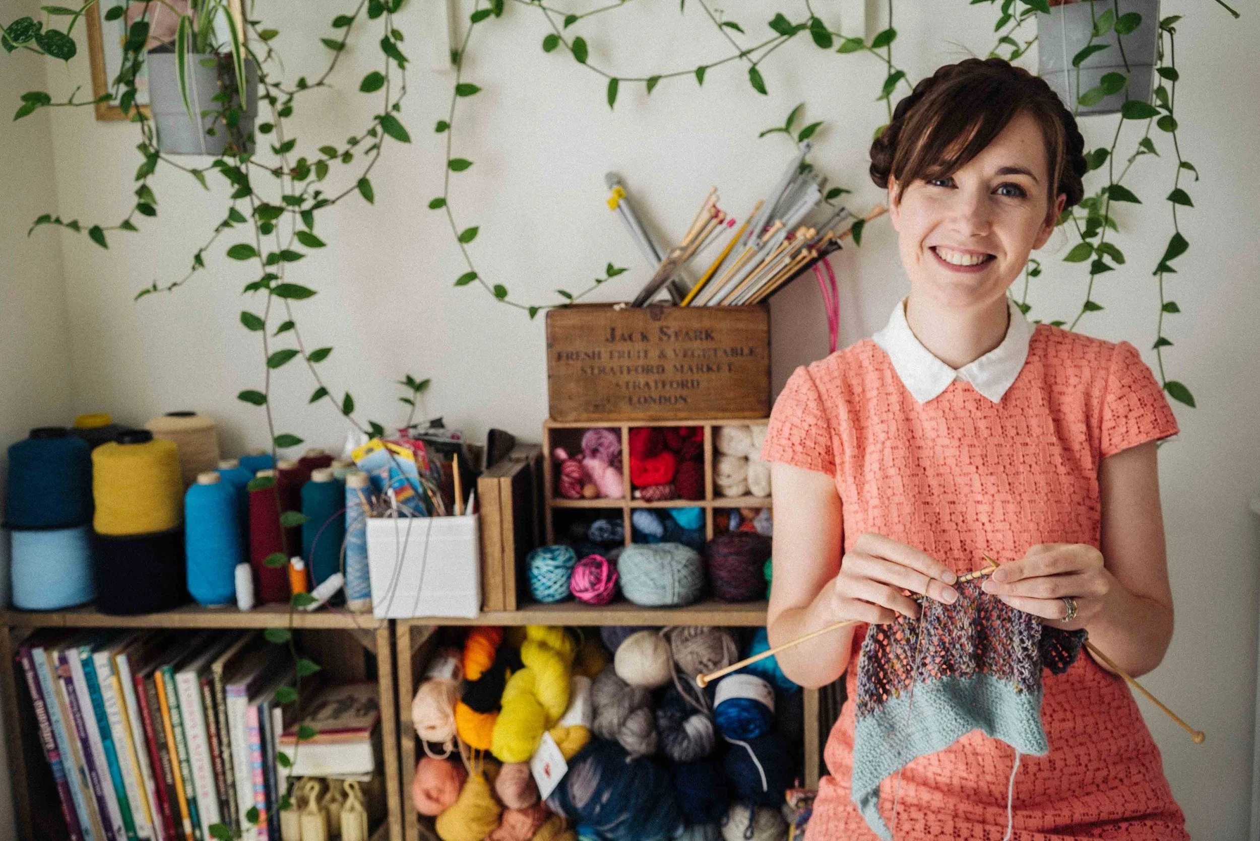 Alice, a white female knitting teacher, sits knitting in a peach dress and white collar.
