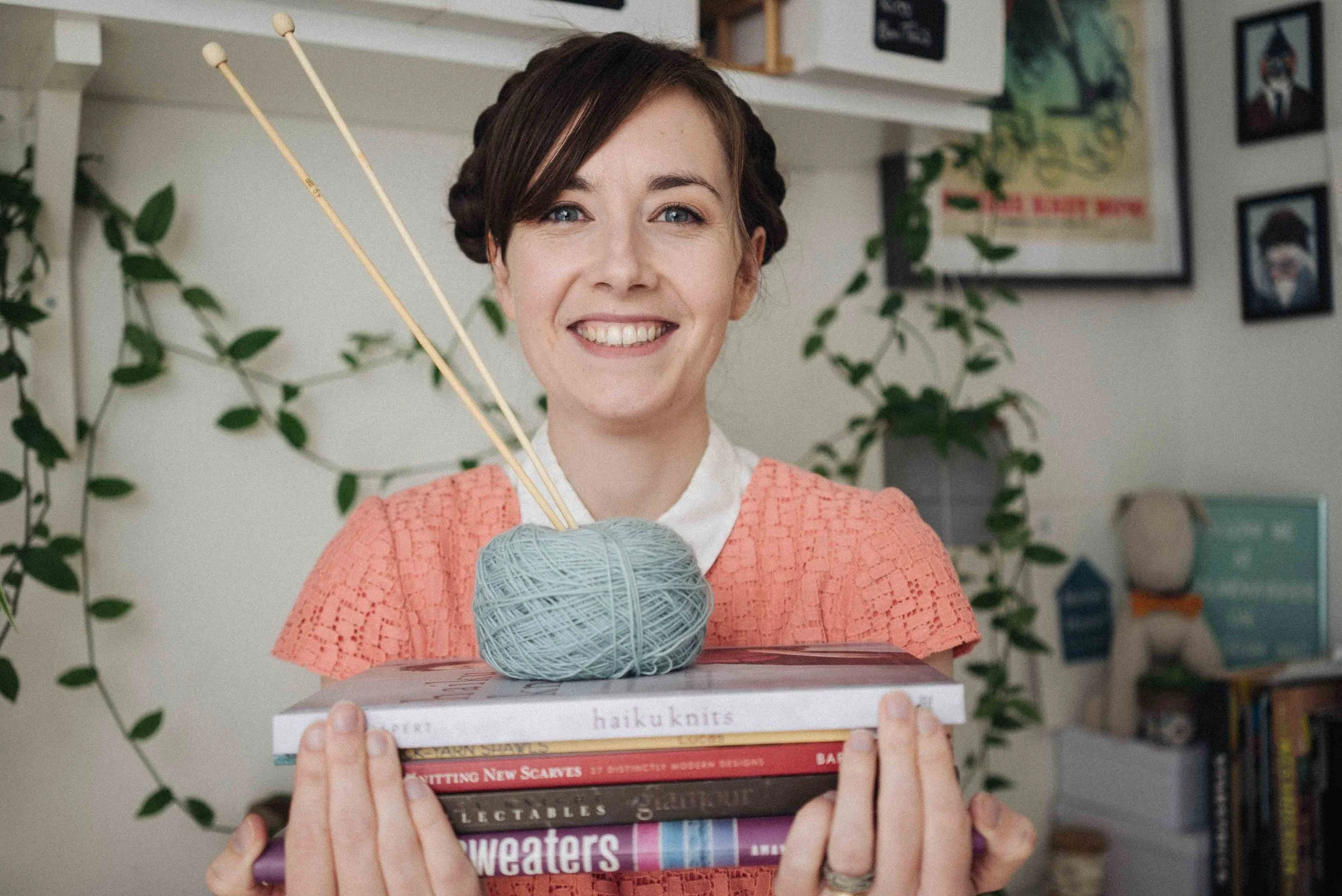 A smiling woman holding a stack of books with knitting pattern books on top, background of houseplants and framed pictures.