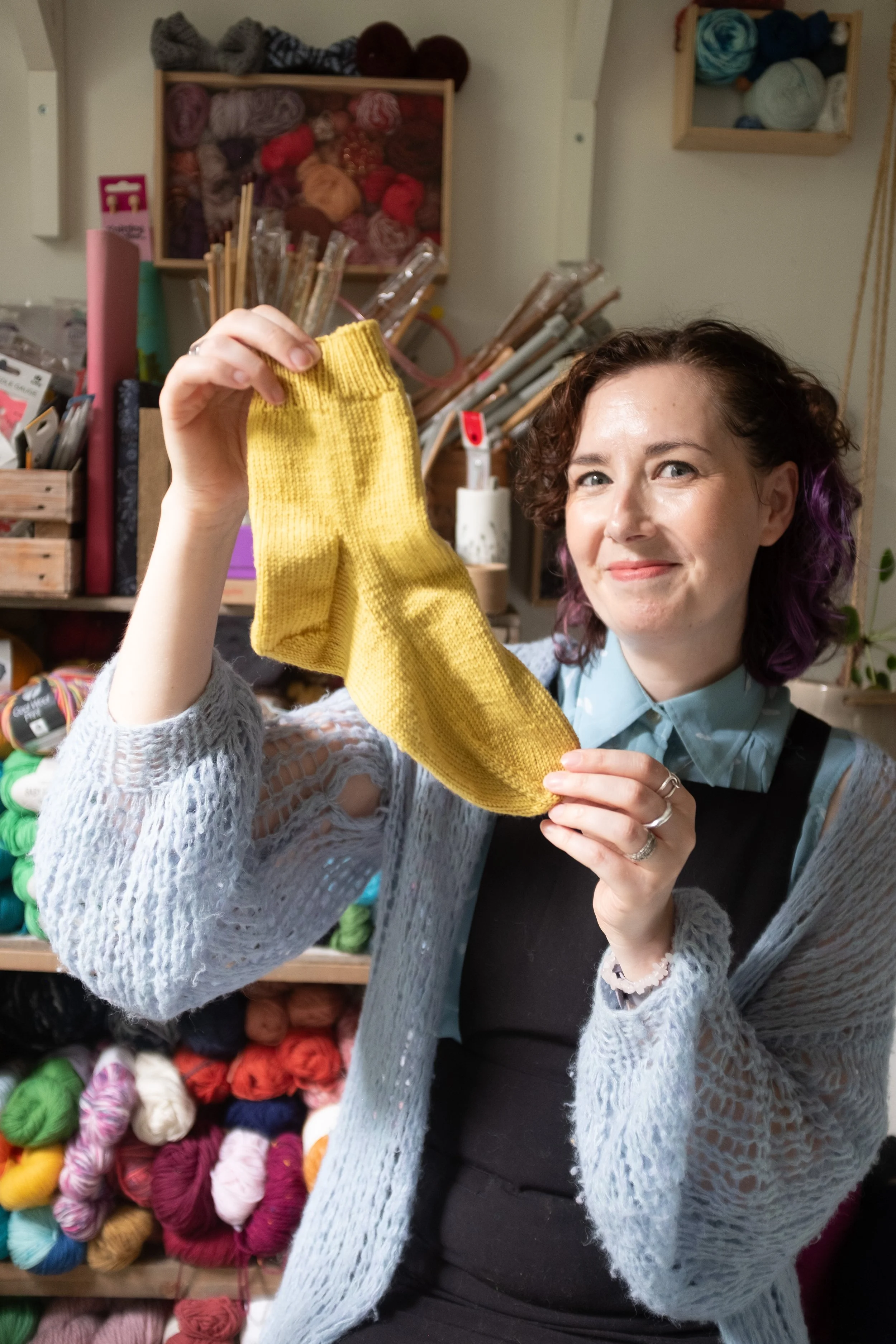 Alice, a white female knitting teacher holds a yellow, handknitted sock and smiles at the camera