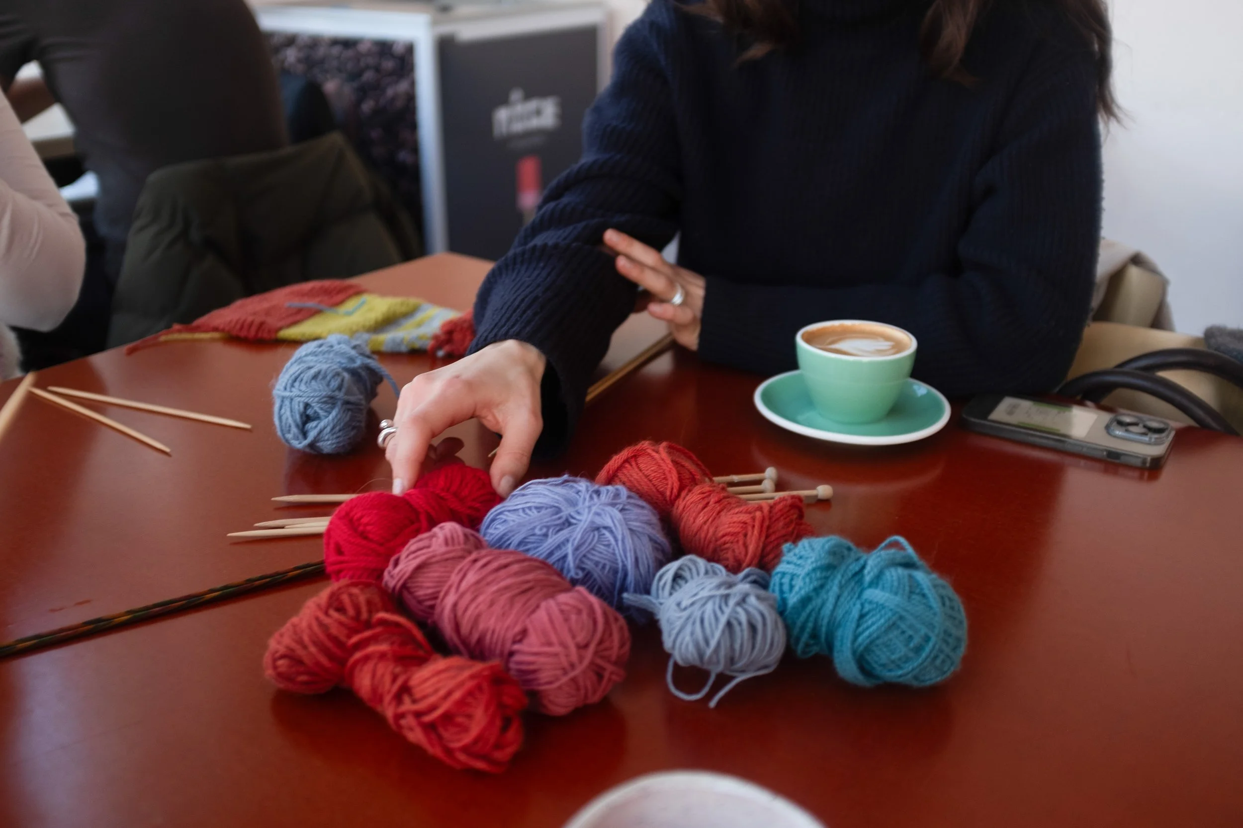 A white female hand reaches for yarn on a table with multiple coloured yarn balls