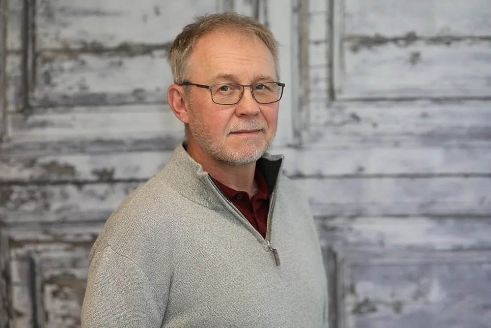 A middle-aged man with gray hair and glasses, wearing a light gray zip-up sweater over a maroon shirt, standing in front of a weathered wooden wall.