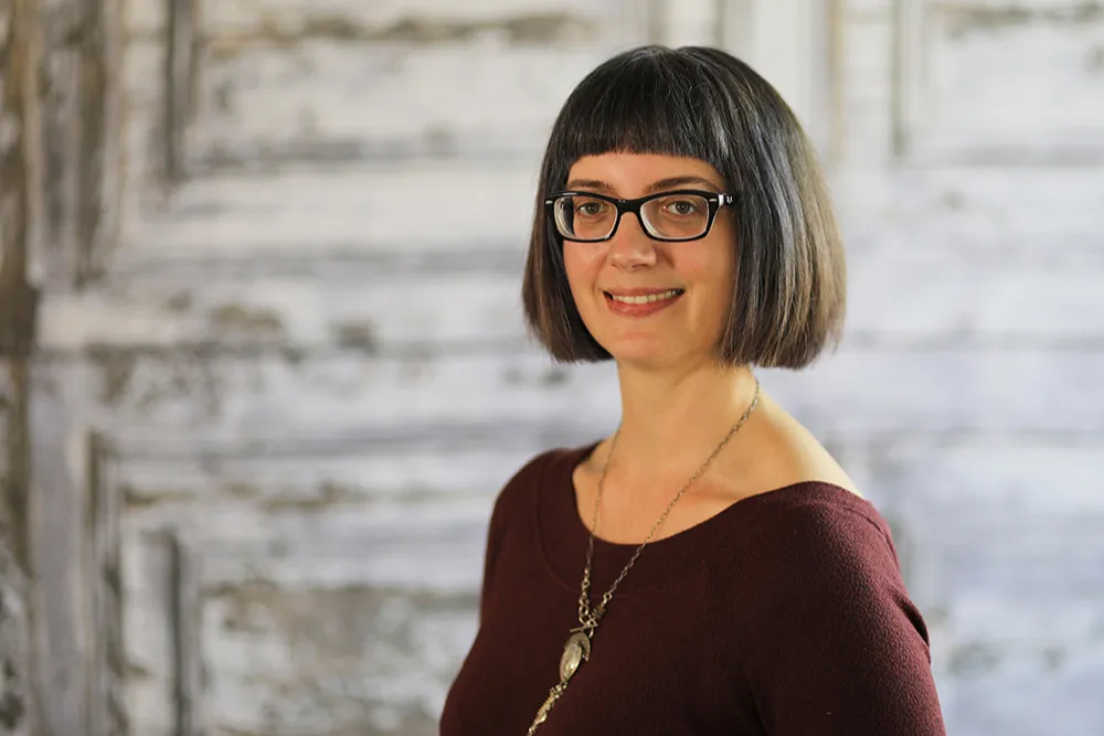 A woman with shoulder-length dark hair, glasses, wearing a burgundy top and a necklace, smiling in front of a rustic wooden background.