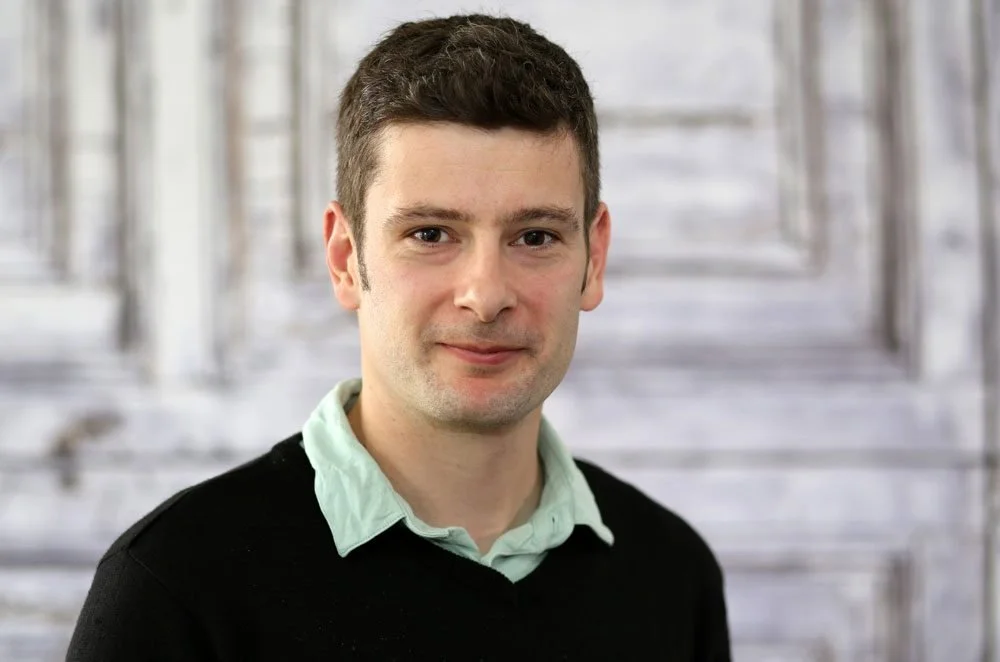 A young man with short dark hair wearing a light green collared shirt and black sweater posing in front of a wooden background.