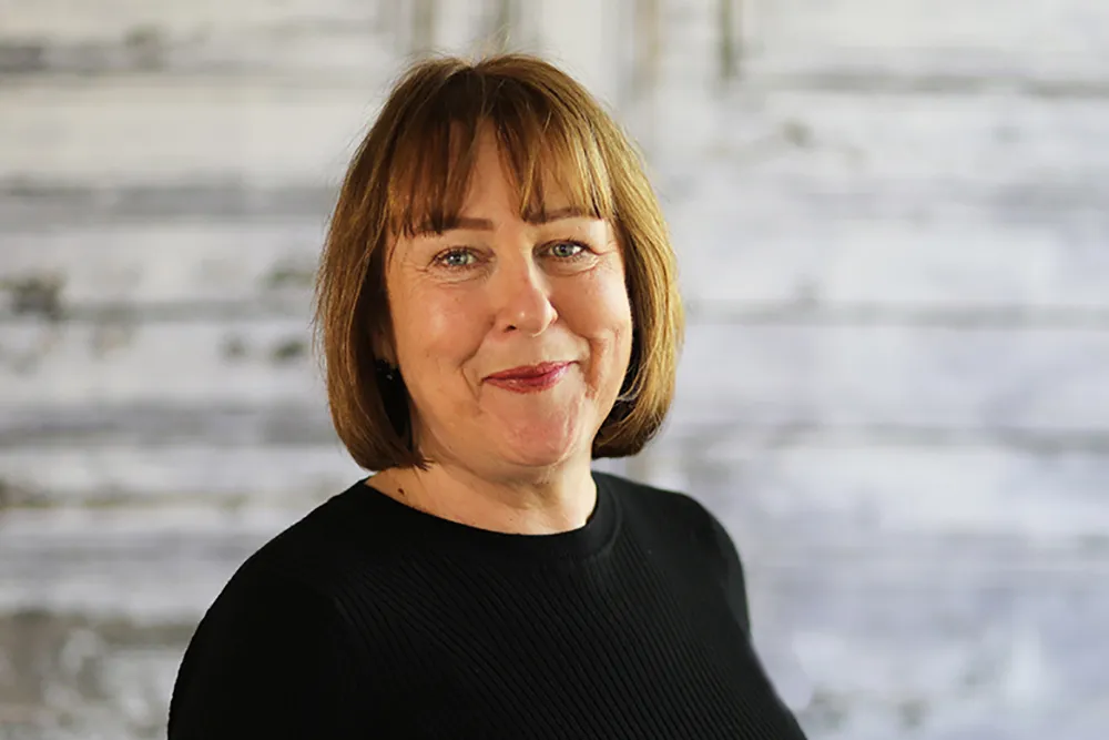 A woman with shoulder-length brown hair wearing a black top, smiling at the camera with a blurred wooden background.