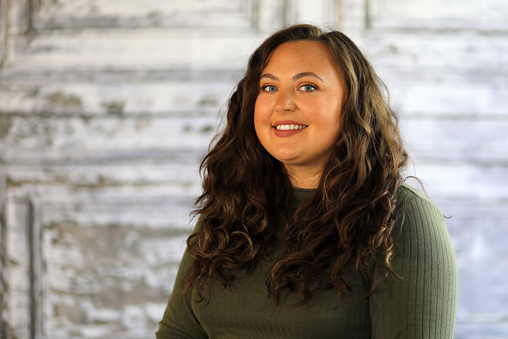 A woman with long, curly brown hair and a green long-sleeve top smiling against a rustic, weathered white wooden background.