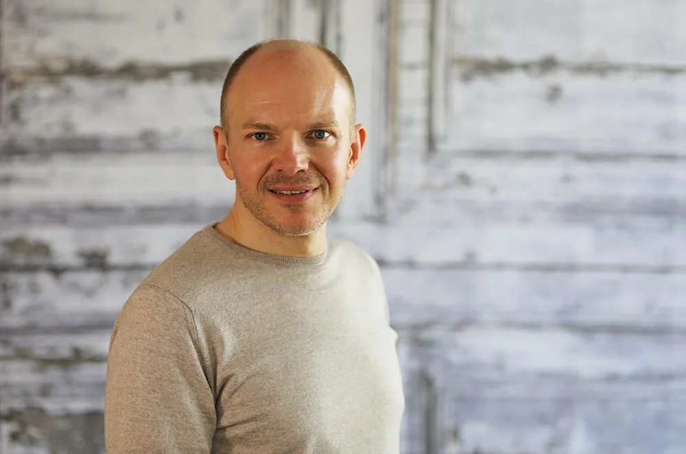 A man with a bald head and light skin wearing a beige sweater smiling at the camera, standing in front of a weathered wood background.