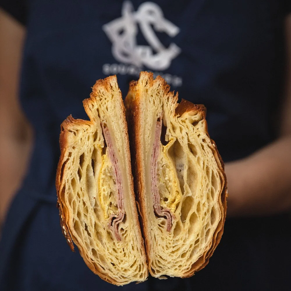 A close-up view of the inside of a sliced croissant sandwich with ham, held by a person wearing a navy shirt with white text and logo.