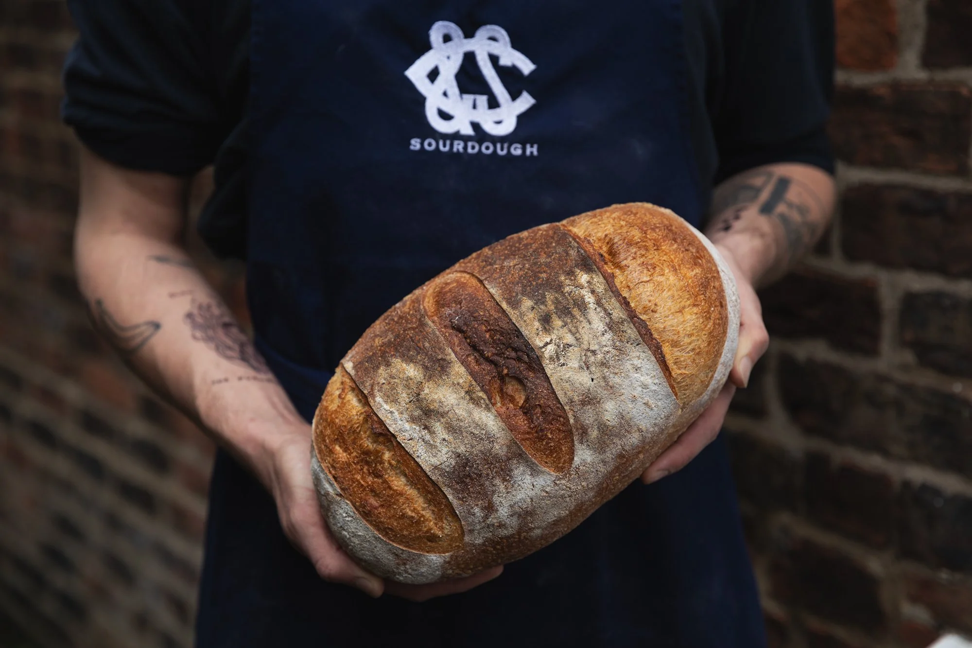 Person wearing a black apron with a bakery logo holding a large loaf of crusty artisan bread in front of a brick wall.