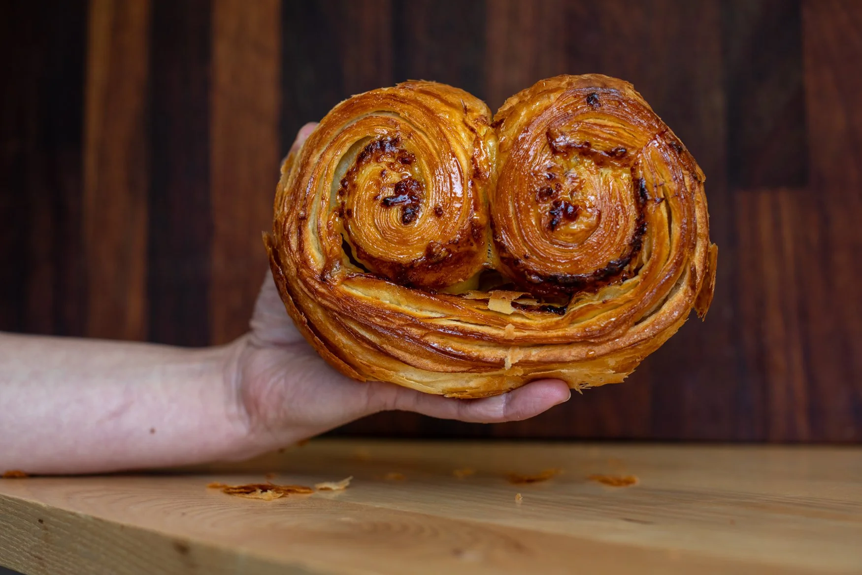 A person holding a large, spiral-shaped pastry with a golden, flaky crust. The pastry appears to be a cinnamon roll or similar baked good, placed over a wooden surface.