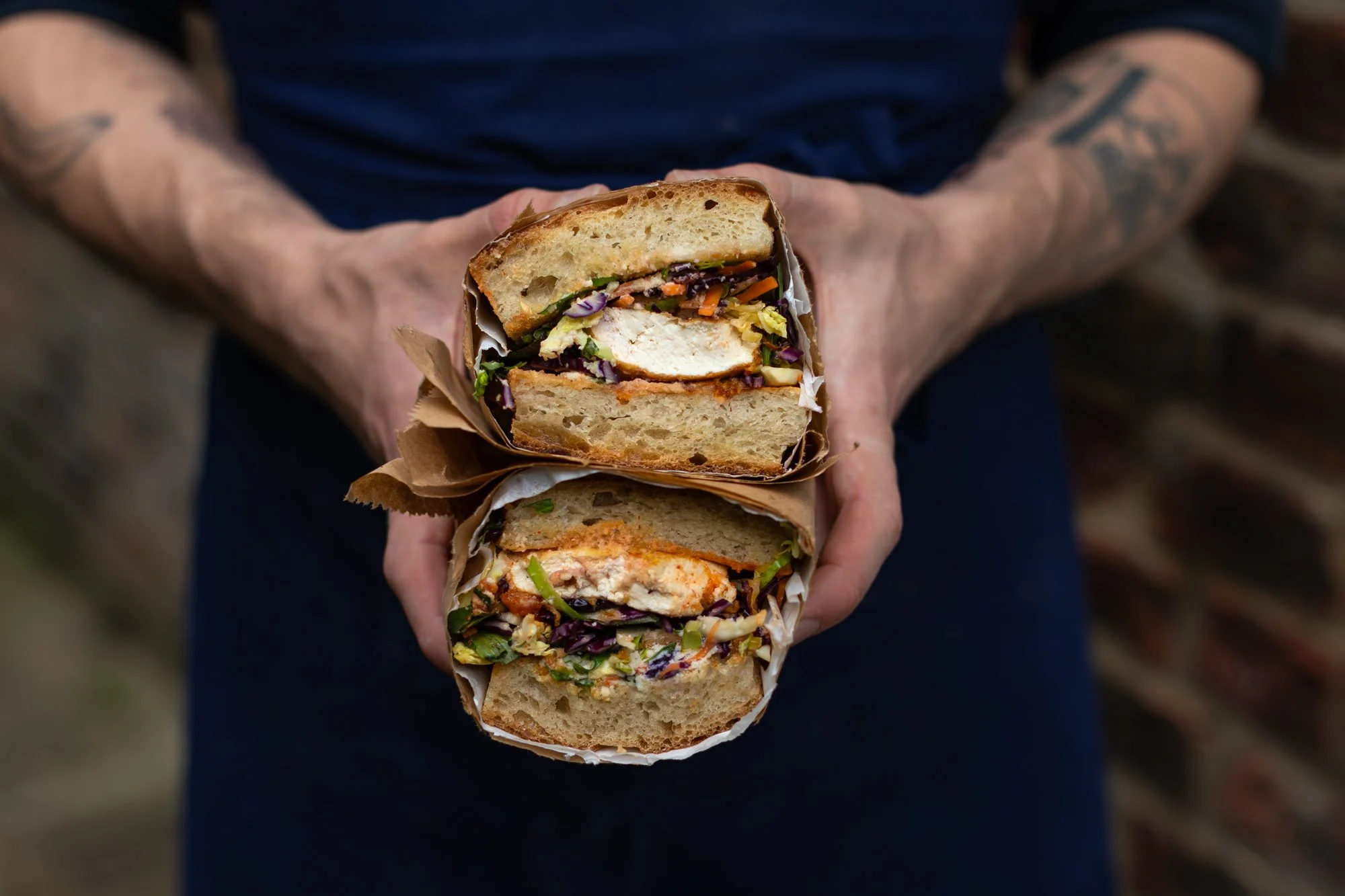 Person holding two halves of a sandwich with visible chicken, lettuce, and other vegetables inside, wrapped in brown paper.