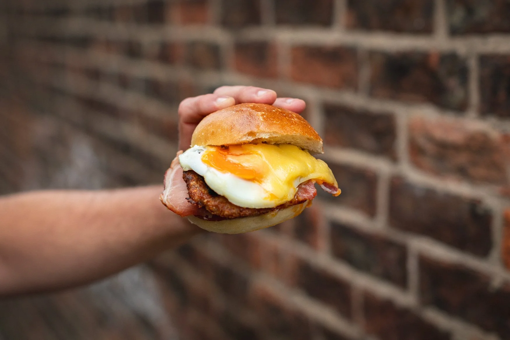 A hand holding a sandwich with a fried egg, bacon, and sausage patty on a bun against a brick wall background.