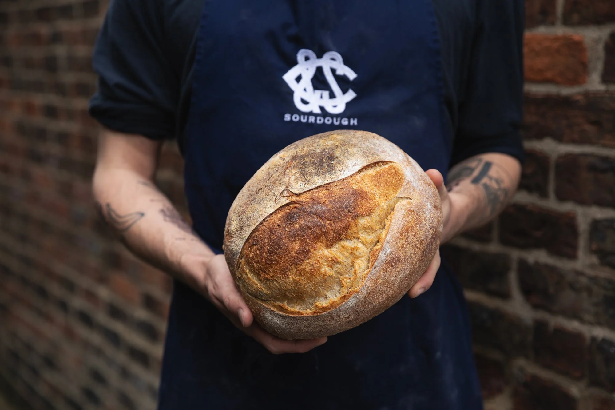 Person holding a round loaf of bread with a crusty top, wearing a dark apron with a sourdough logo, standing against a brick wall.