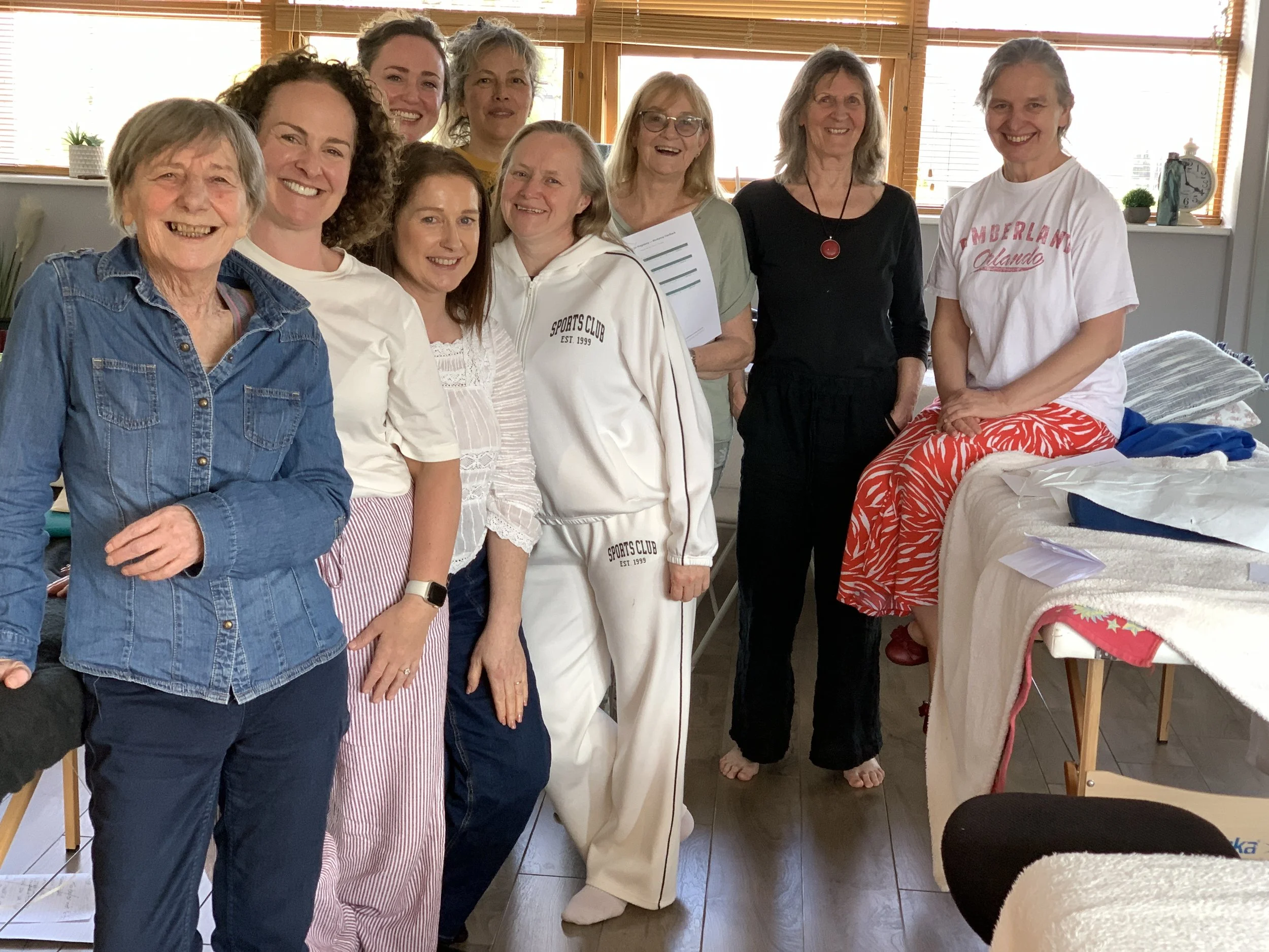 Group of ten women standing together indoors, smiling for a photo. They are dressed casually, with a window and blinds in the background, and various household items visible.