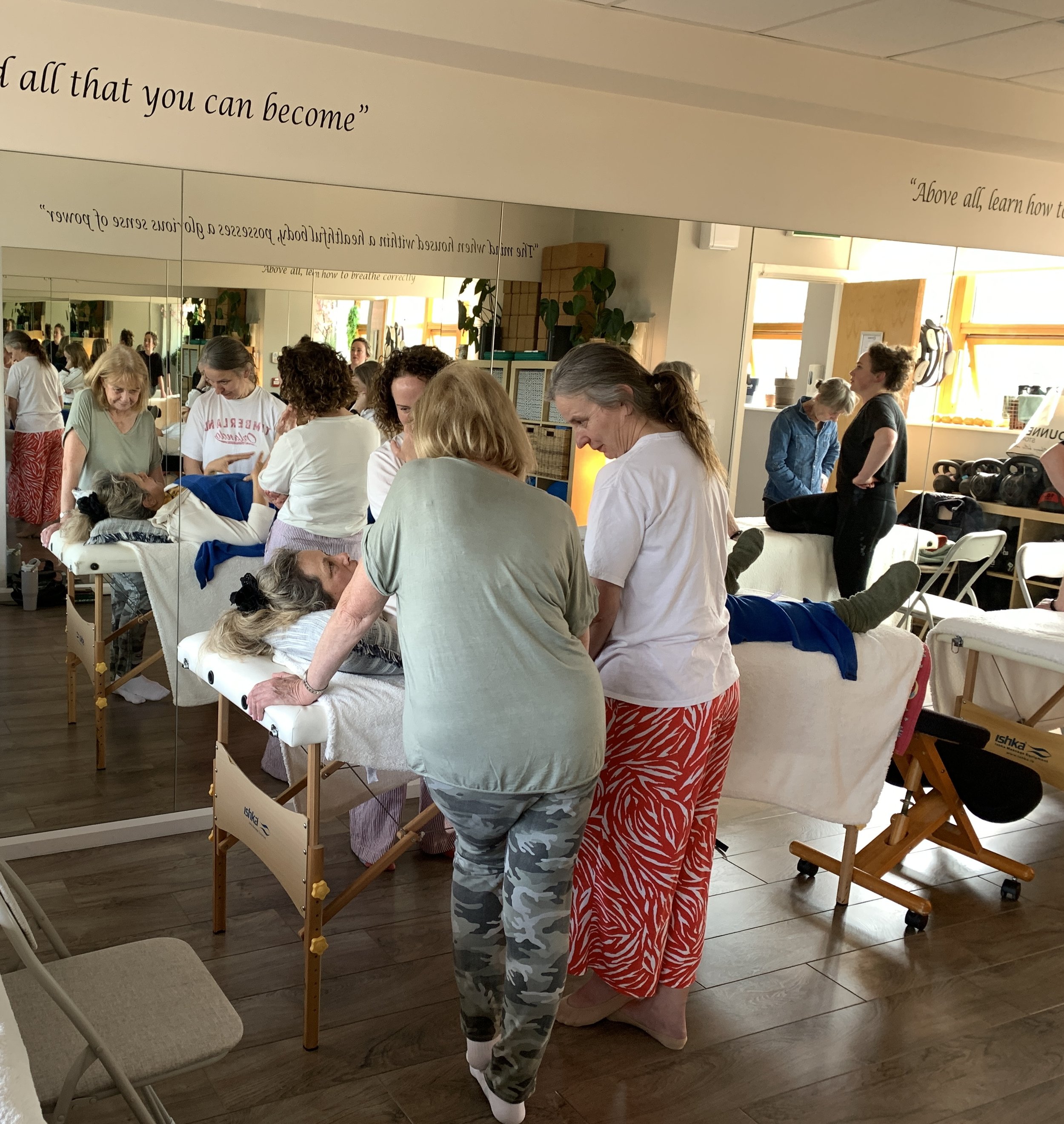 A group of women gathered around a massage table in a wellness or therapy center, with some women performing or receiving treatments. The room has sunlight coming through windows, and there are various chairs, plants, and therapy equipment visible.