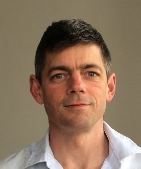 A headshot of a man with short dark hair, fair skin, and wearing a white collared shirt, against a plain neutral background.