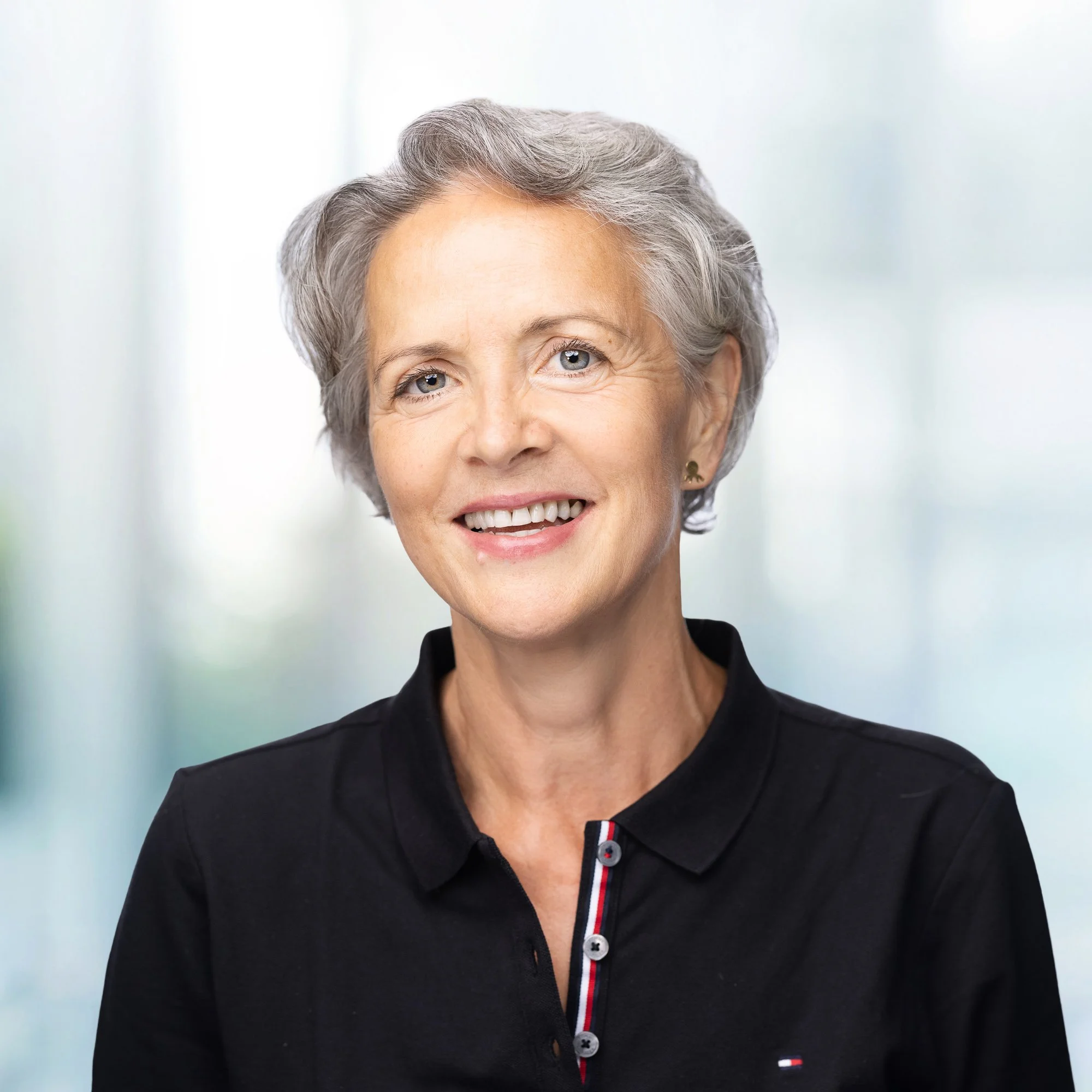 Close-up of an older woman with short gray hair, blue eyes, and a bright smile, wearing a black polo shirt with a small logo and a striped collar, standing in front of a blurred light-colored background.