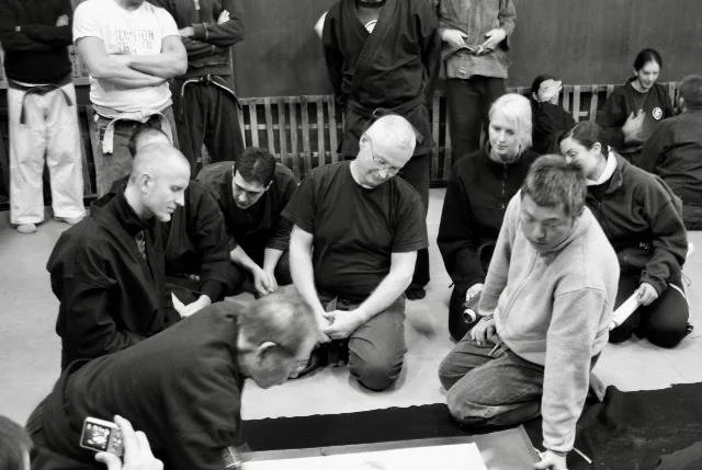 A group of people gathered in a dojo or training room, with some kneeling or sitting on the floor, watching a martial arts demonstration.