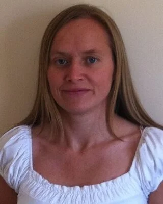 A woman with long blonde hair wearing a white top with puffy short sleeves, standing against a plain wall.