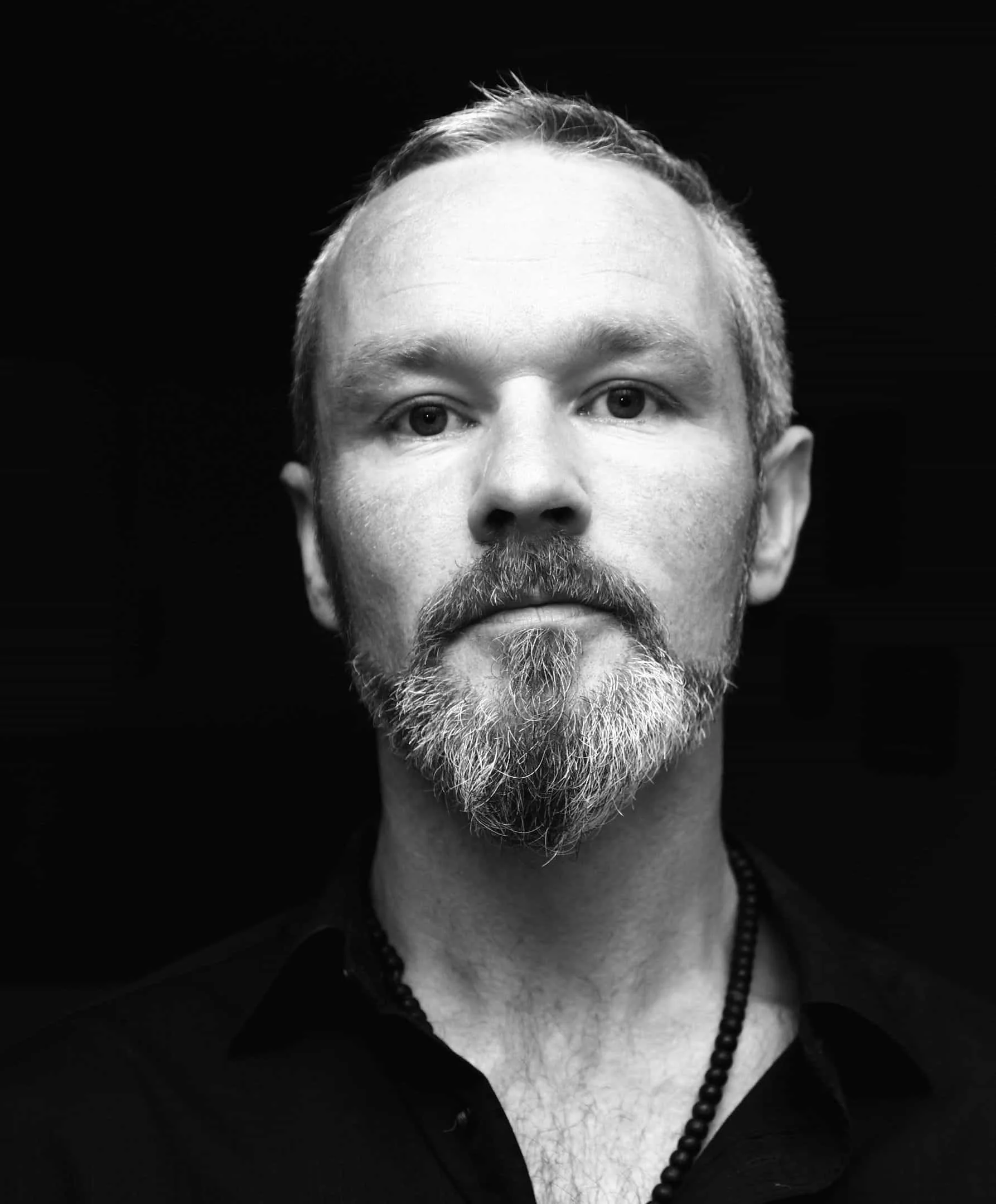 Black and white portrait of a middle-aged man with short hair, beard, and mustache, wearing a dark shirt and beaded necklace.