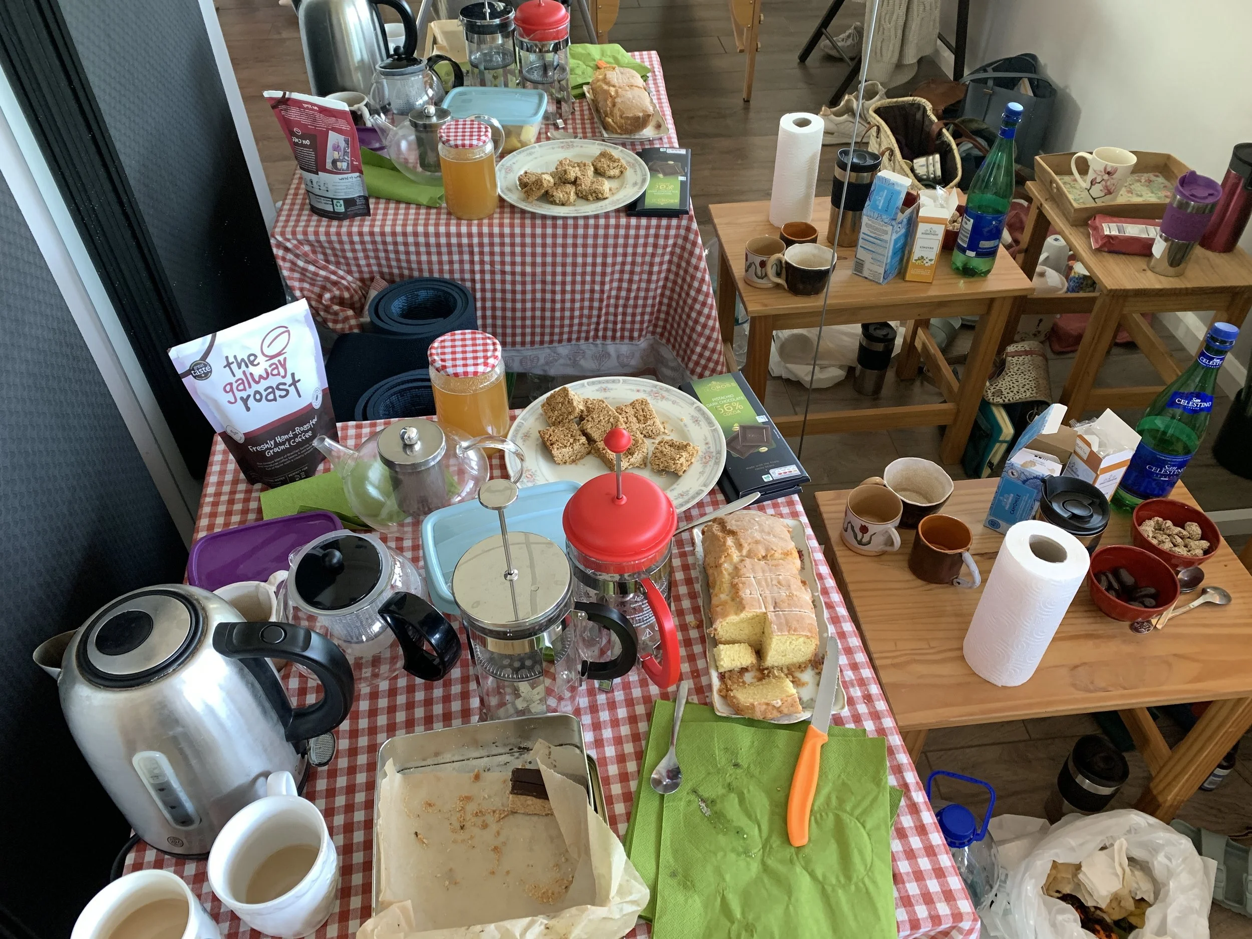 A cluttered kitchen table with a red and white checkered tablecloth, holding various food items including a loaf of bread, granola bars, bowls, a teapot, cups, juice, and utensils, with kitchen items and supplies scattered around.