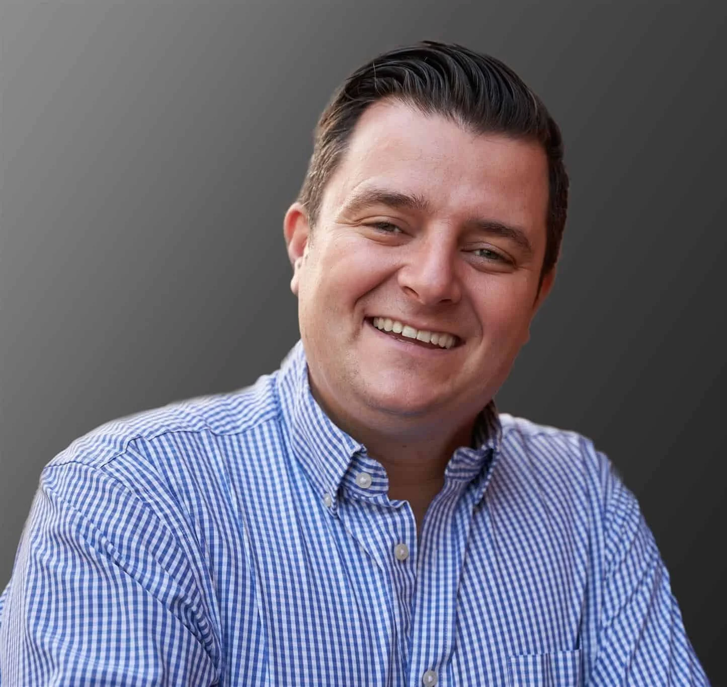 A smiling man wearing a blue checkered shirt, looking at the camera against a plain gray background.