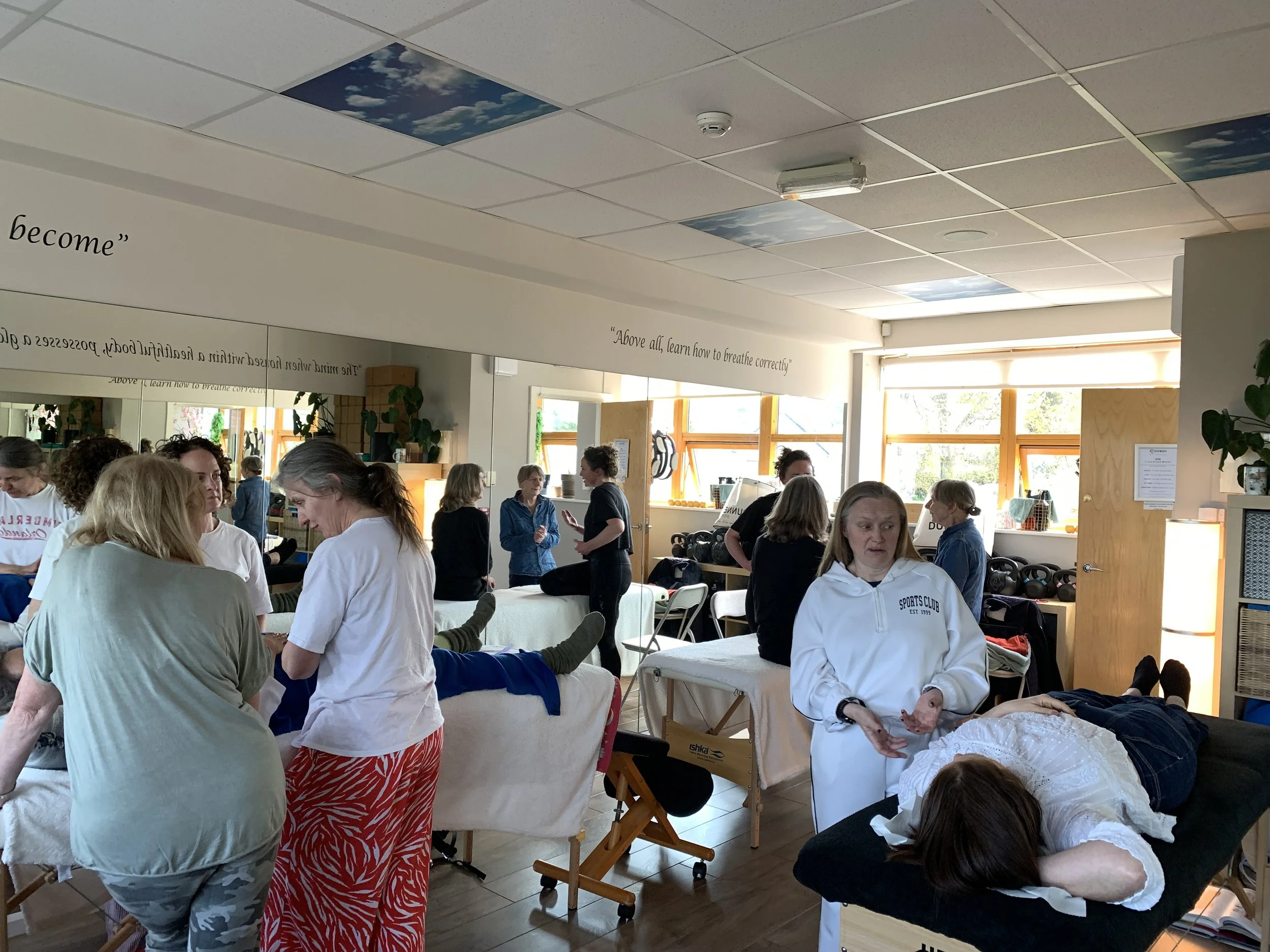 Several people engaged in a wellness or massage session in a bright room with large windows. Some are receiving treatment on massage tables, while others are standing around and talking. The room has ceiling tiles with images of clouds and sky, and wall decorations with quotes about breathing and becoming.