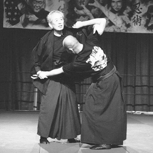 Two martial artists practicing Aikido on stage, one appears to be demonstrating a technique while the other is falling backward.