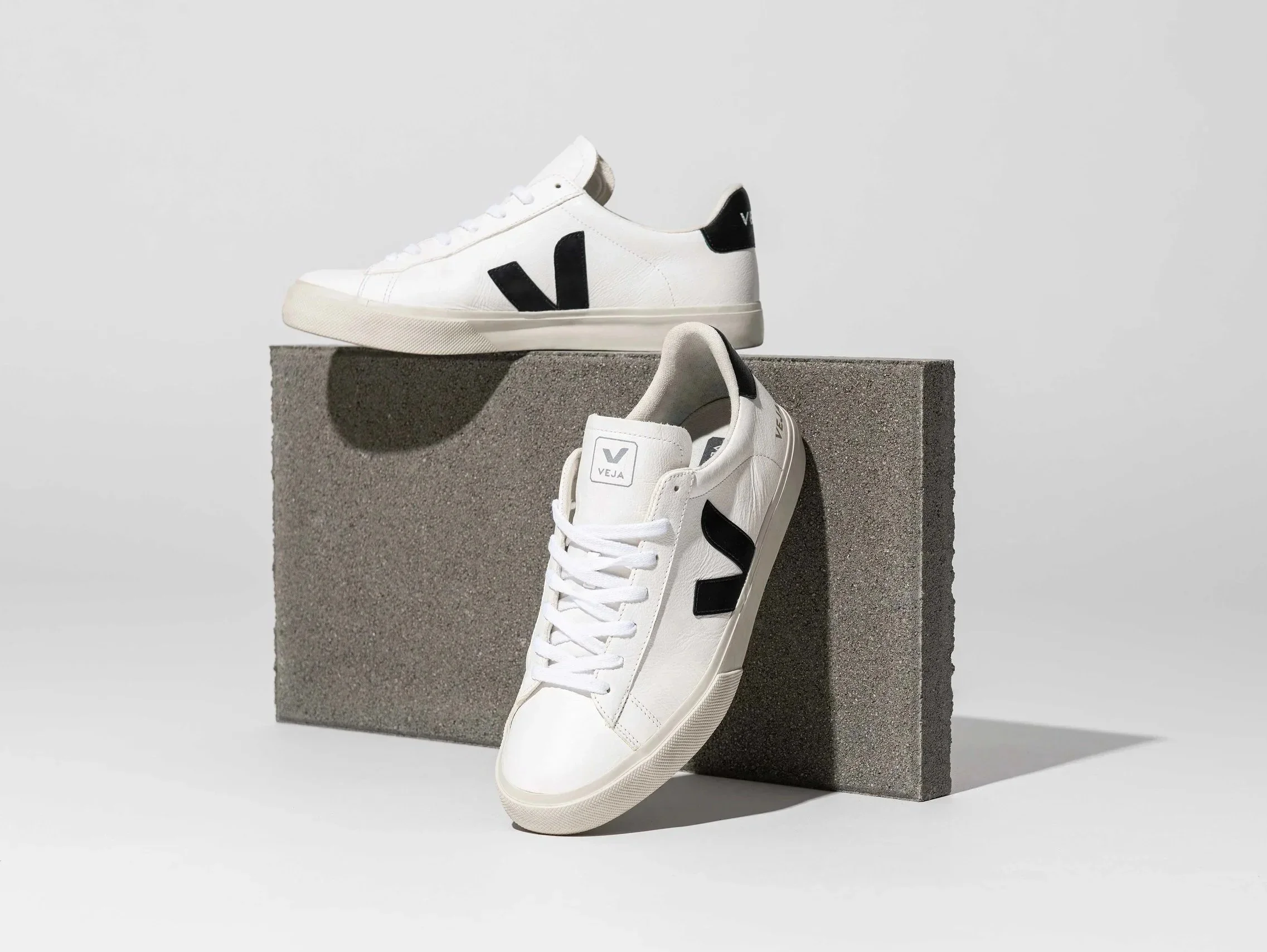 Two white sneakers with black logo accents are displayed, one shoe leaning against a gray stone block and the other resting on the block, against a plain light gray background.