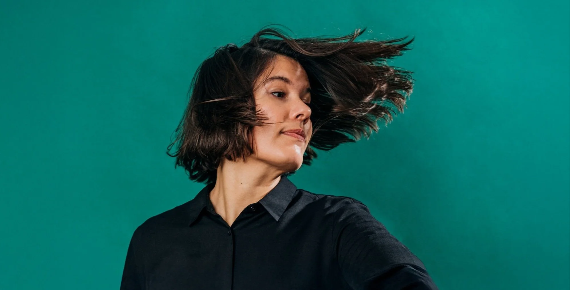 Woman with dark hair, wearing a black shirt, tossing her hair back against a solid green background.