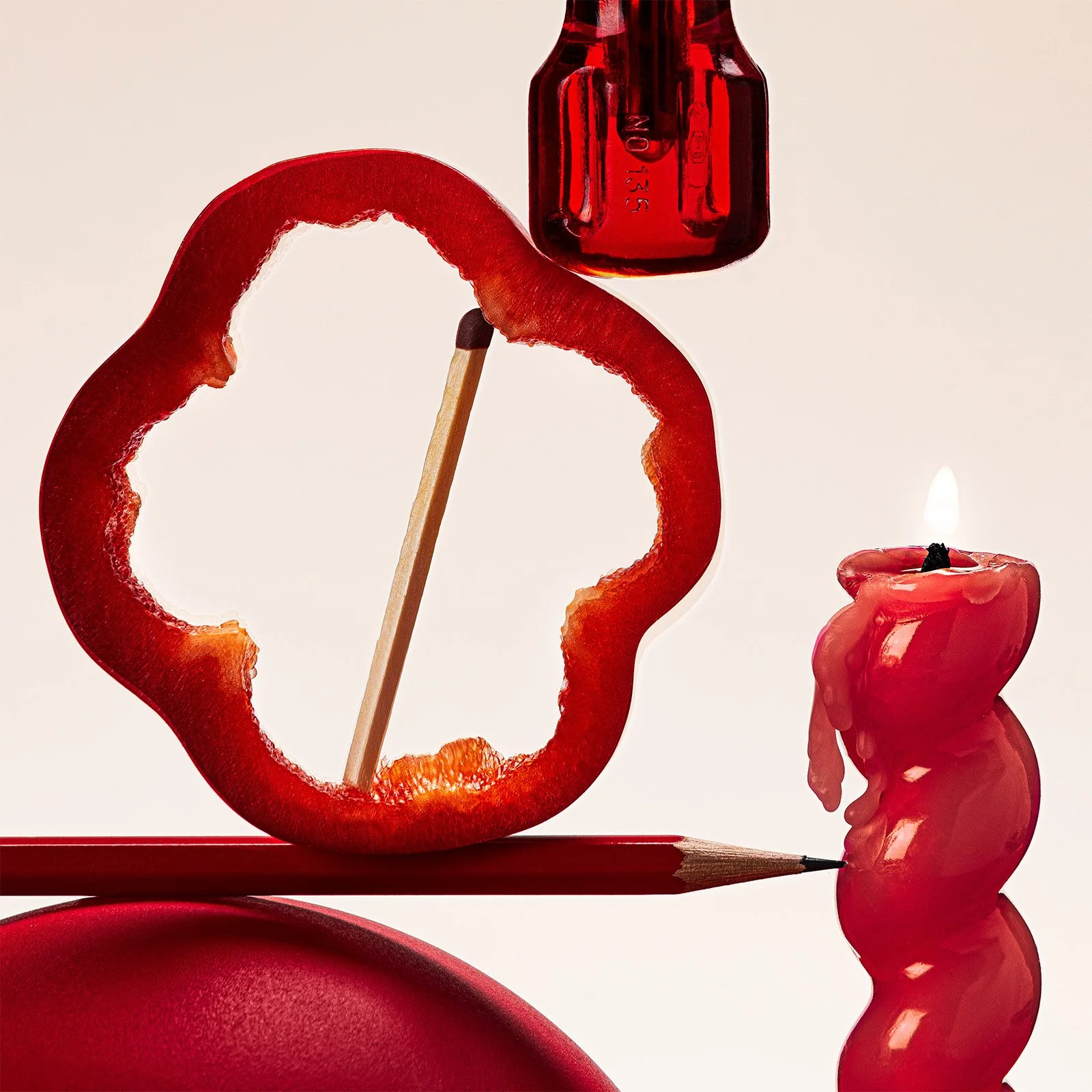 Close-up of sliced red bell pepper, a red glass bottle, a red wax candle, and a red pencil on a white background.