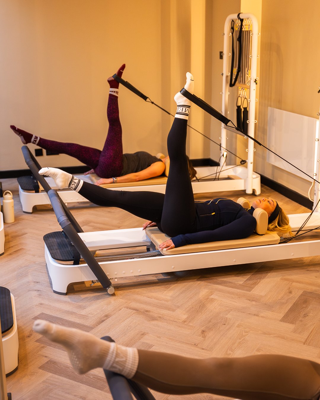 Two women doing pilates exercises on reformer machines in a fitness studio. They are lying on their backs with one leg raised and connected to a resistance band. Both women are wearing workout clothes and socks.