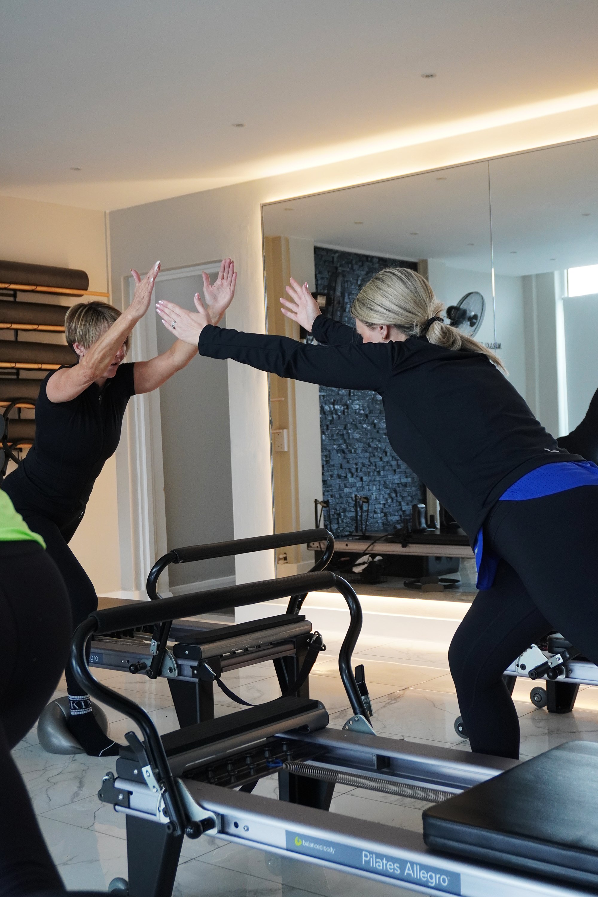 Two women practicing Pilates in a studio, performing a stretching exercise using a Pilates reformer machine.