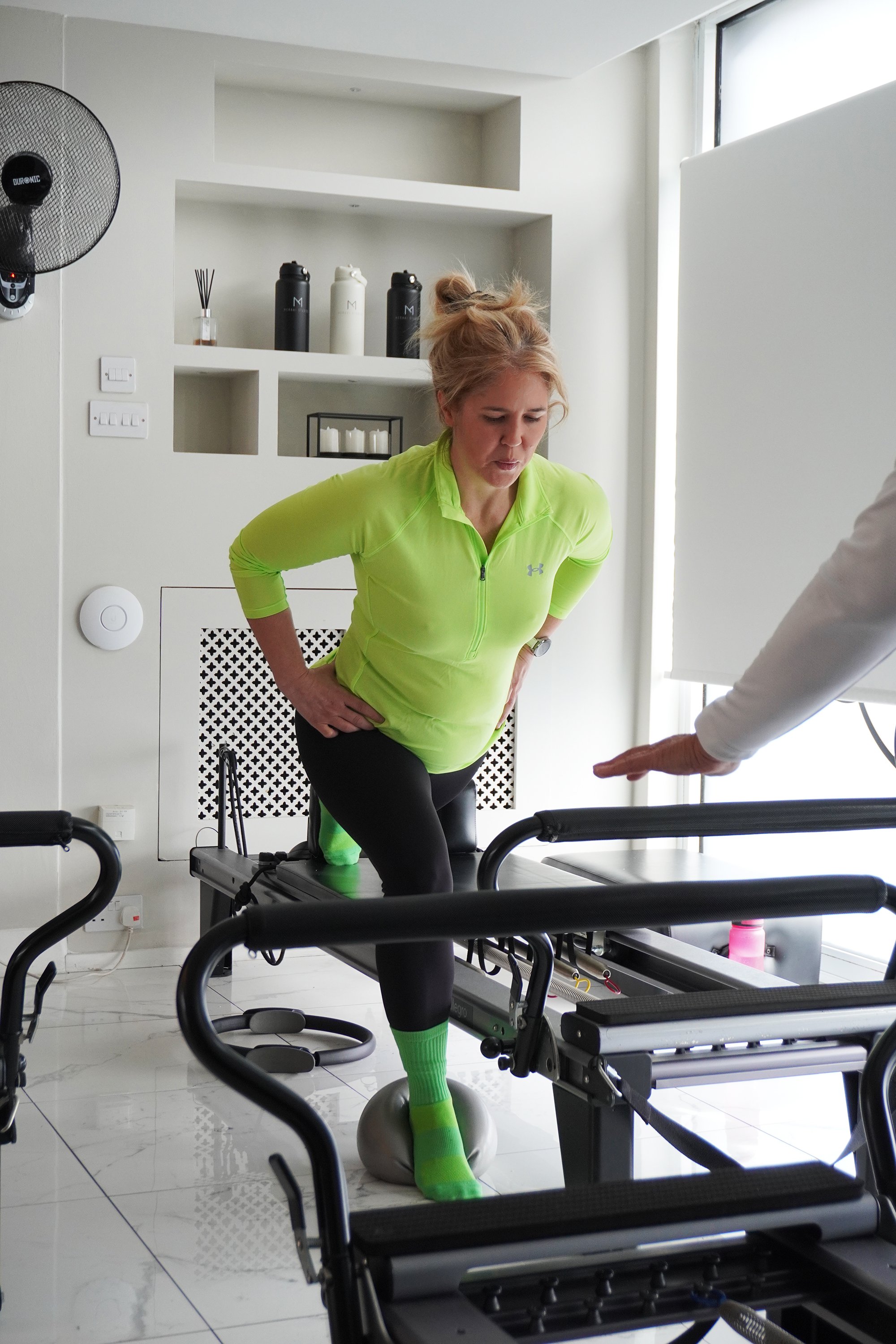 Woman in neon green athletic clothing performing a rehab exercise on a treadmill with a physiotherapist guiding her, in a bright, modern workout space.