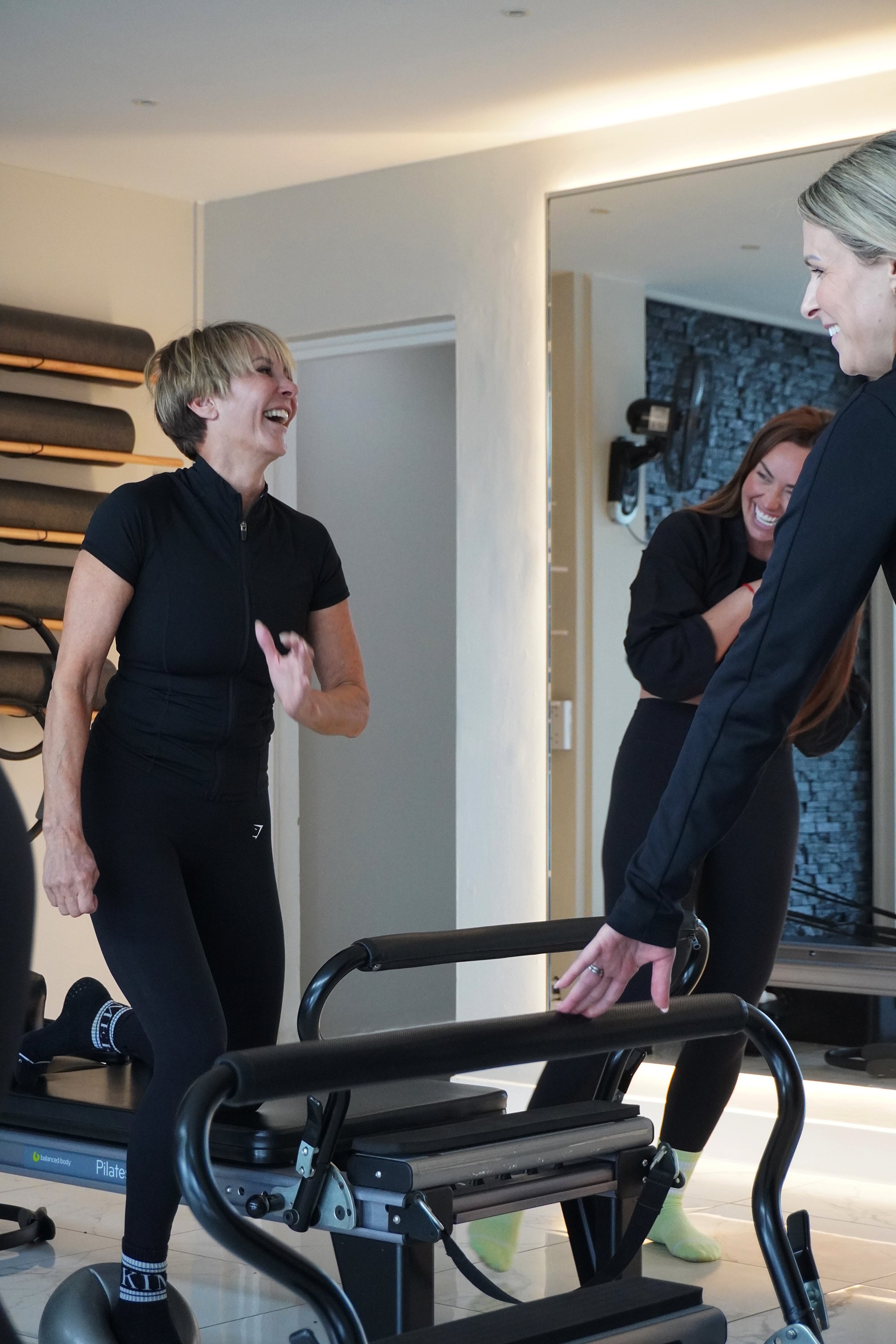 Three women in black workout clothing laughing and talking while in a fitness studio, with Pilates equipment and a mirror in the background.