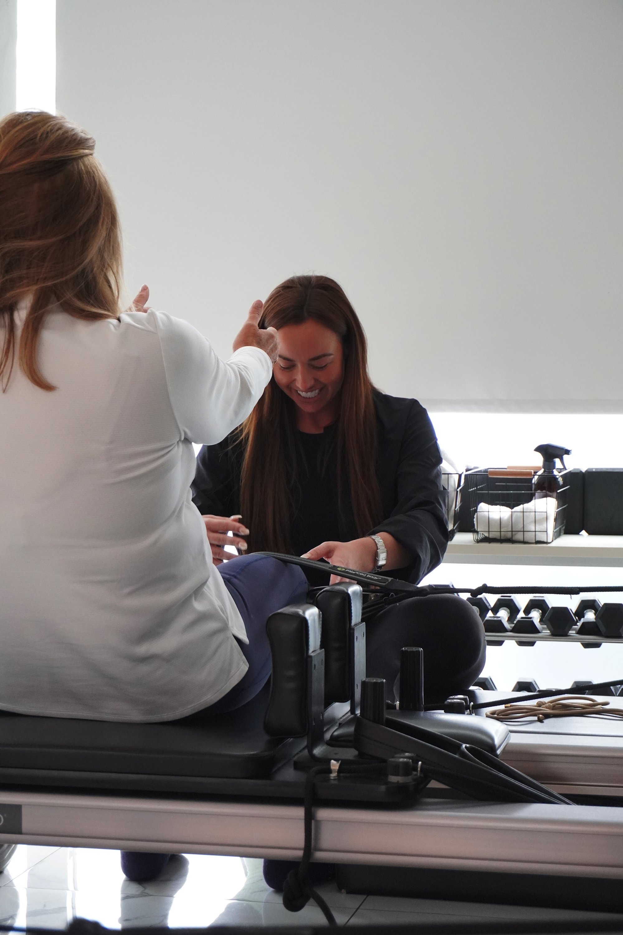A woman sitting on a physical therapy or rehabilitation table, smiling and looking down as a therapist or trainer assists her, in a clinical or therapy setting.