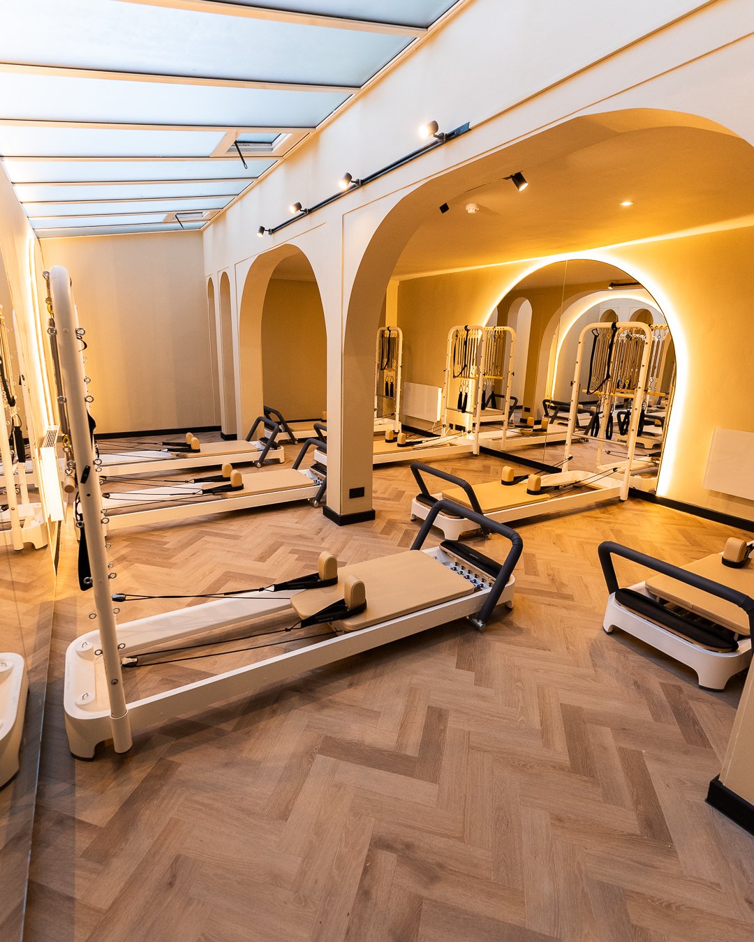 Fitness studio with Pilates reformer machines, mirrors, and wooden flooring, illuminated by soft lighting and skylights.