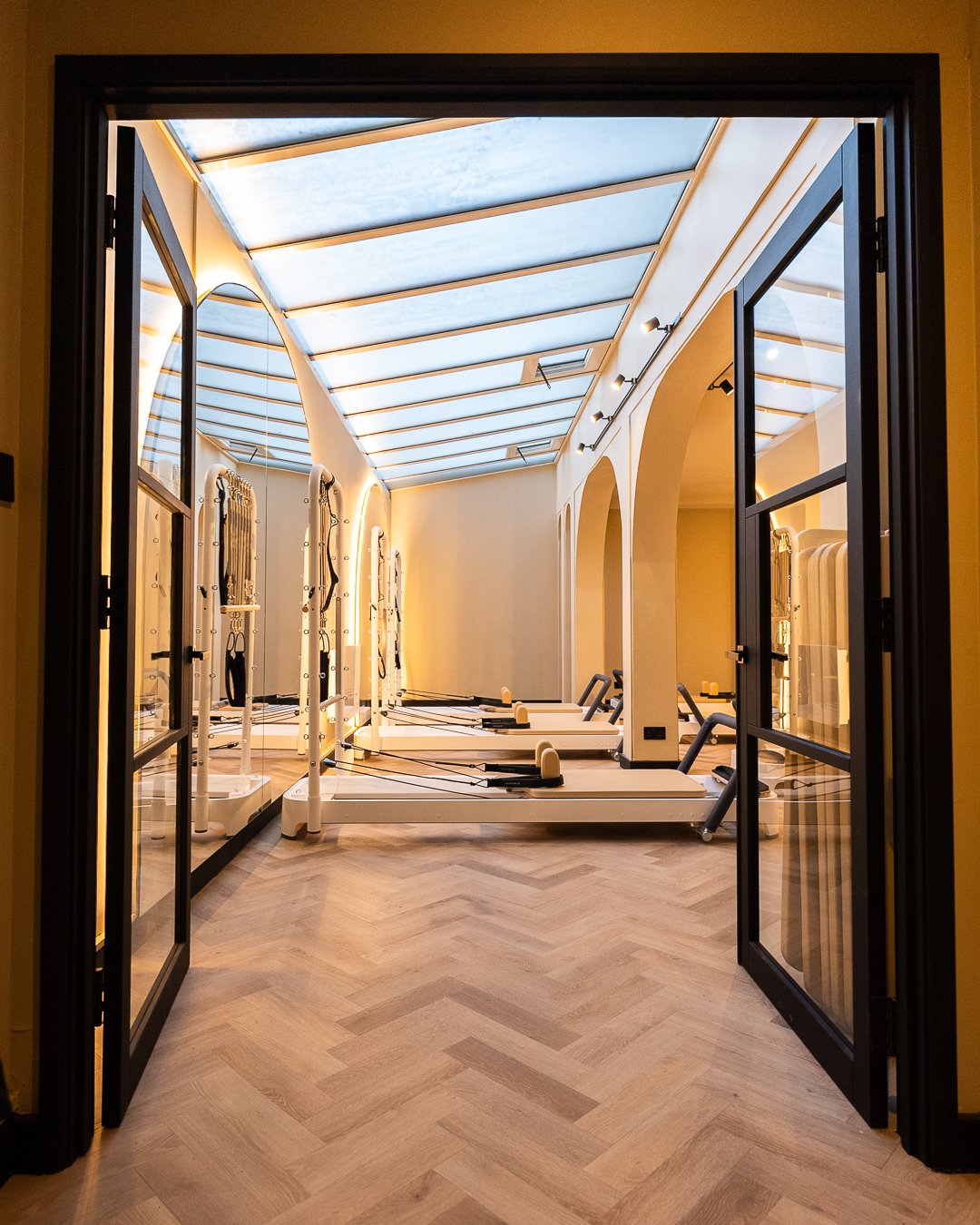 A bright room with a glass ceiling, showcasing multiple Pilates reformer machines lined up against the wall, seen through open double glass doors.