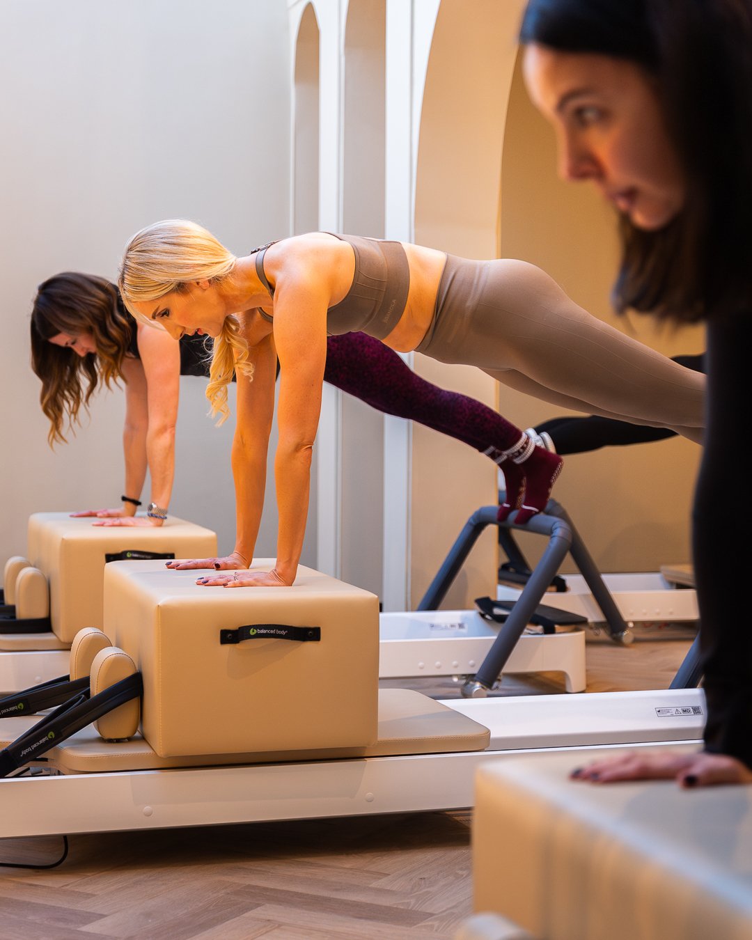Three women in athletic attire doing reformer Pilates exercises on Pilates reformer machines in a studio, with an instructor assisting.