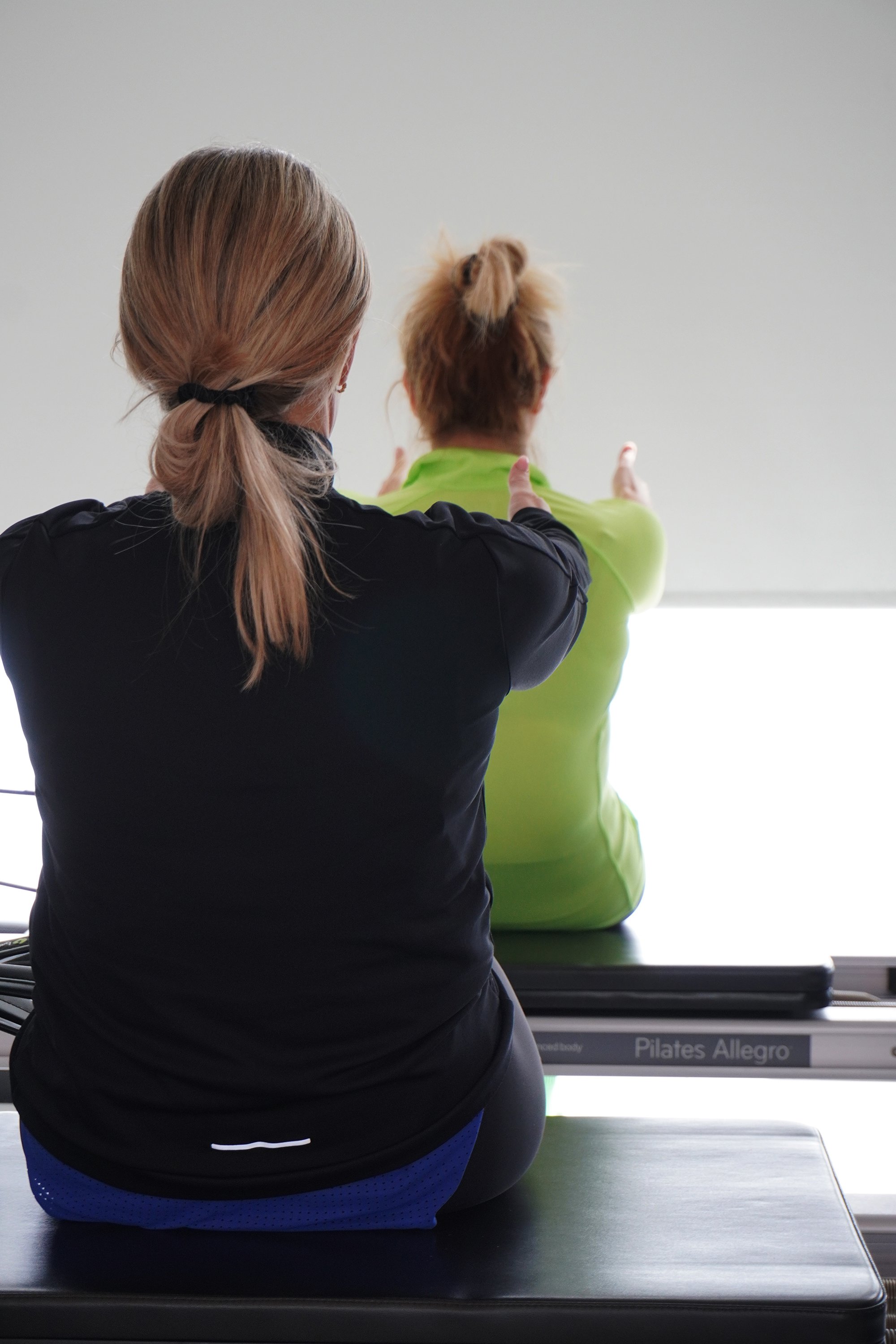 A woman in black athleticwear assists another woman in a green jacket with an exercise or stretching motion during a fitness class or training session.